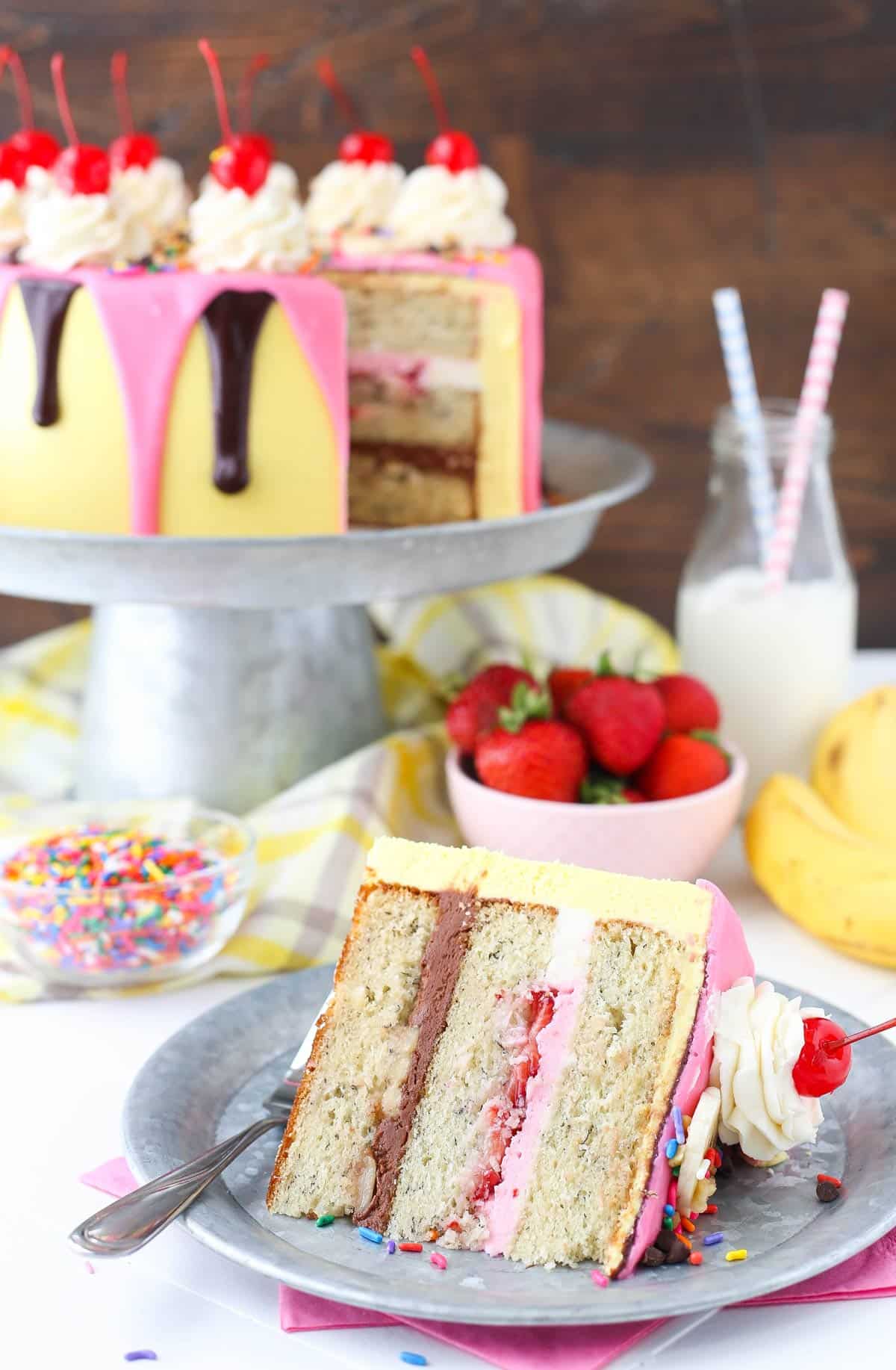 A slice of banana split layer cake with the rest of the cake in the background.
