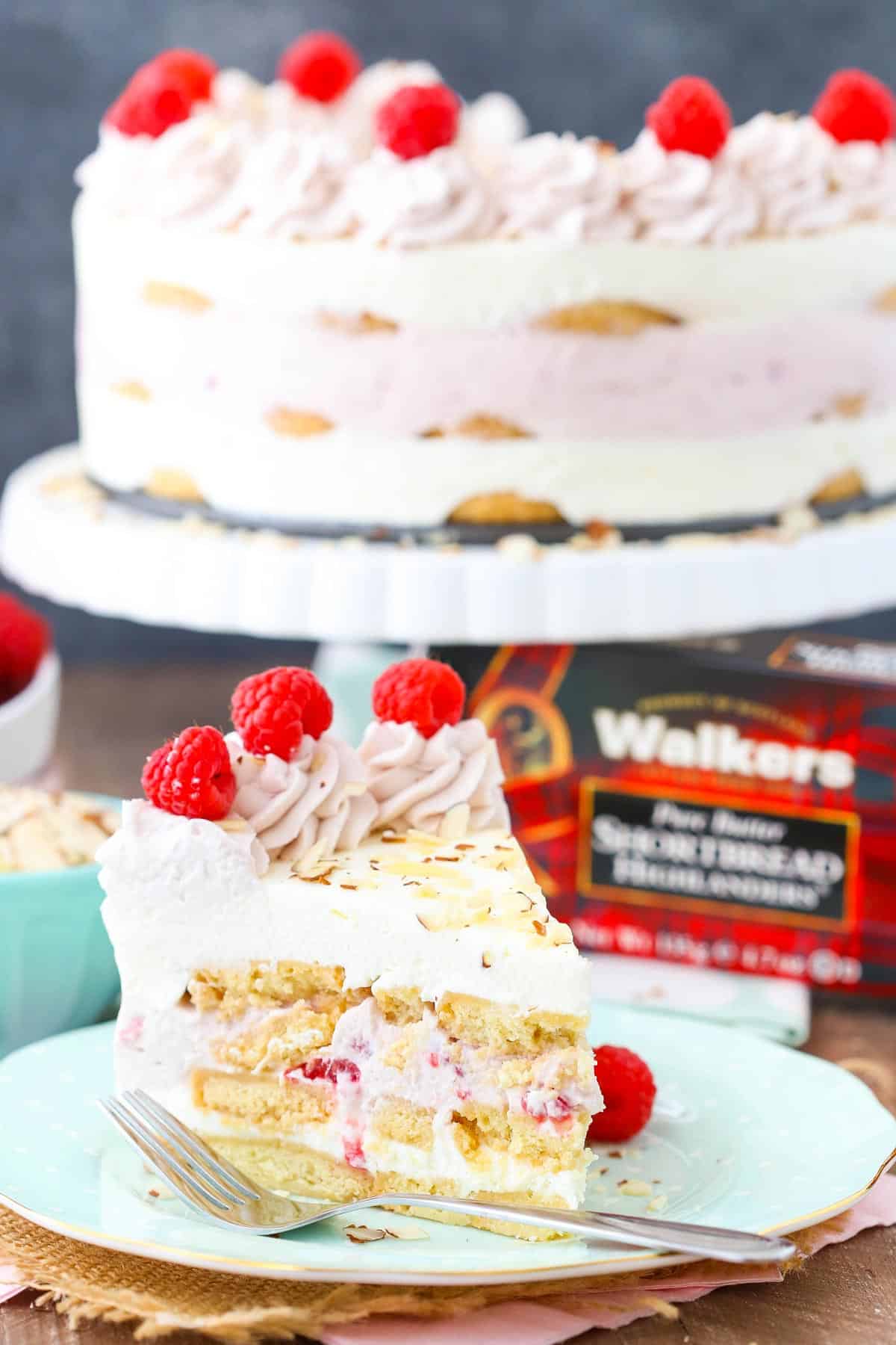 Side view of a slice of raspberry almond shortbread icebox cake on a light blue plate with the rest of the cake in the background.
