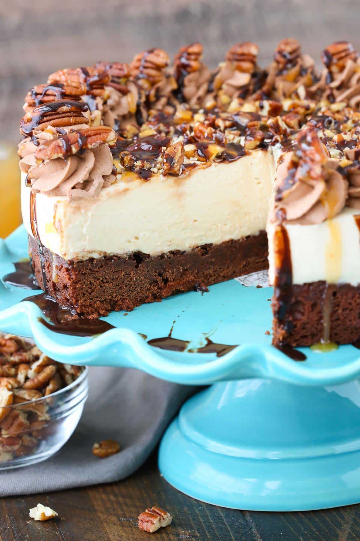A turtle brownie cheesecake on a blue cake stand with a slice removed to show layers.