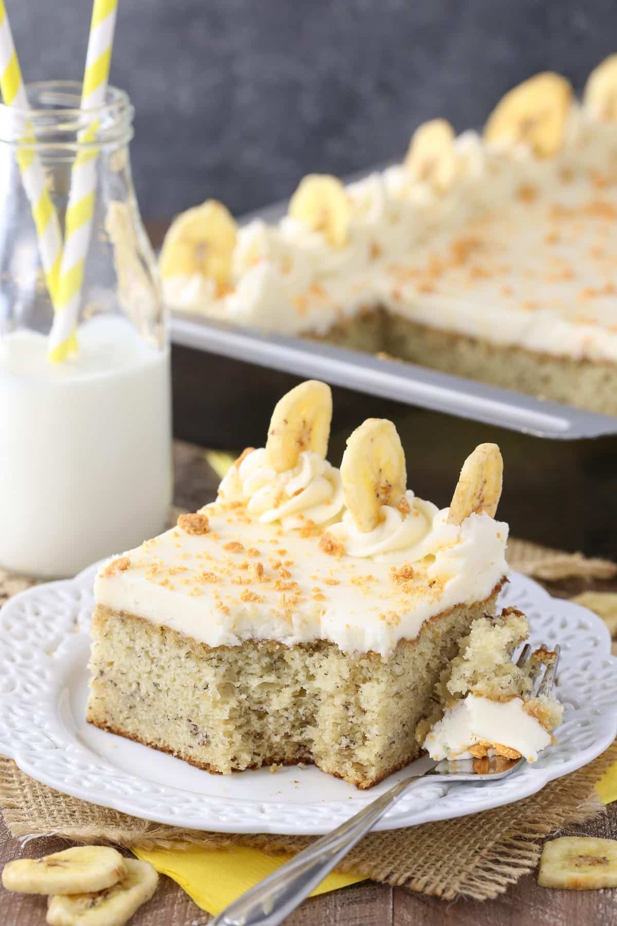 A slice of banana cake with cream cheese frosting on a white plate with a bite removed on a fork.