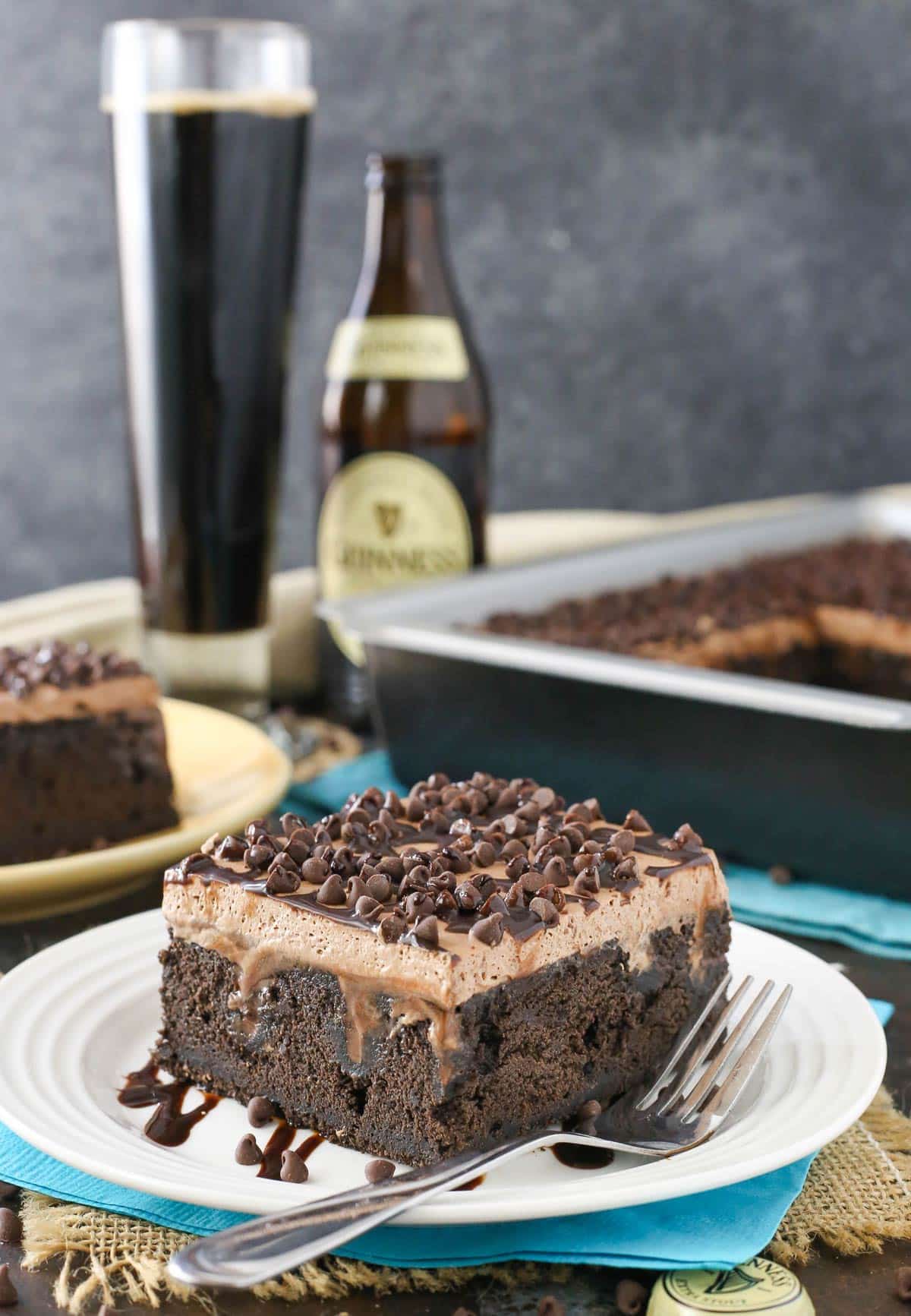 A slice of Guinness chocolate poke cake on a white plate with the rest of the cake in the background.