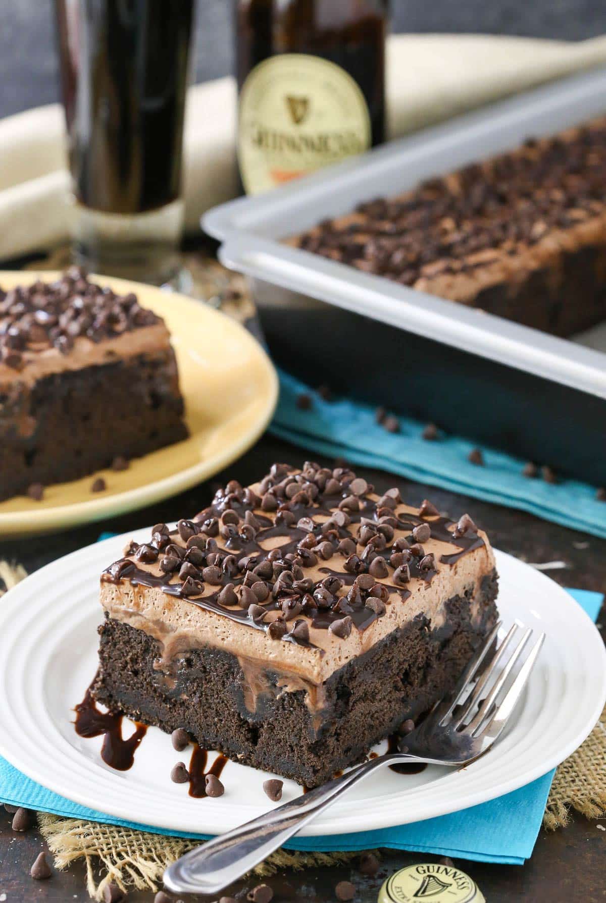 A slice of Guinness chocolate poke cake on a white plate with the rest of the cake in the background.