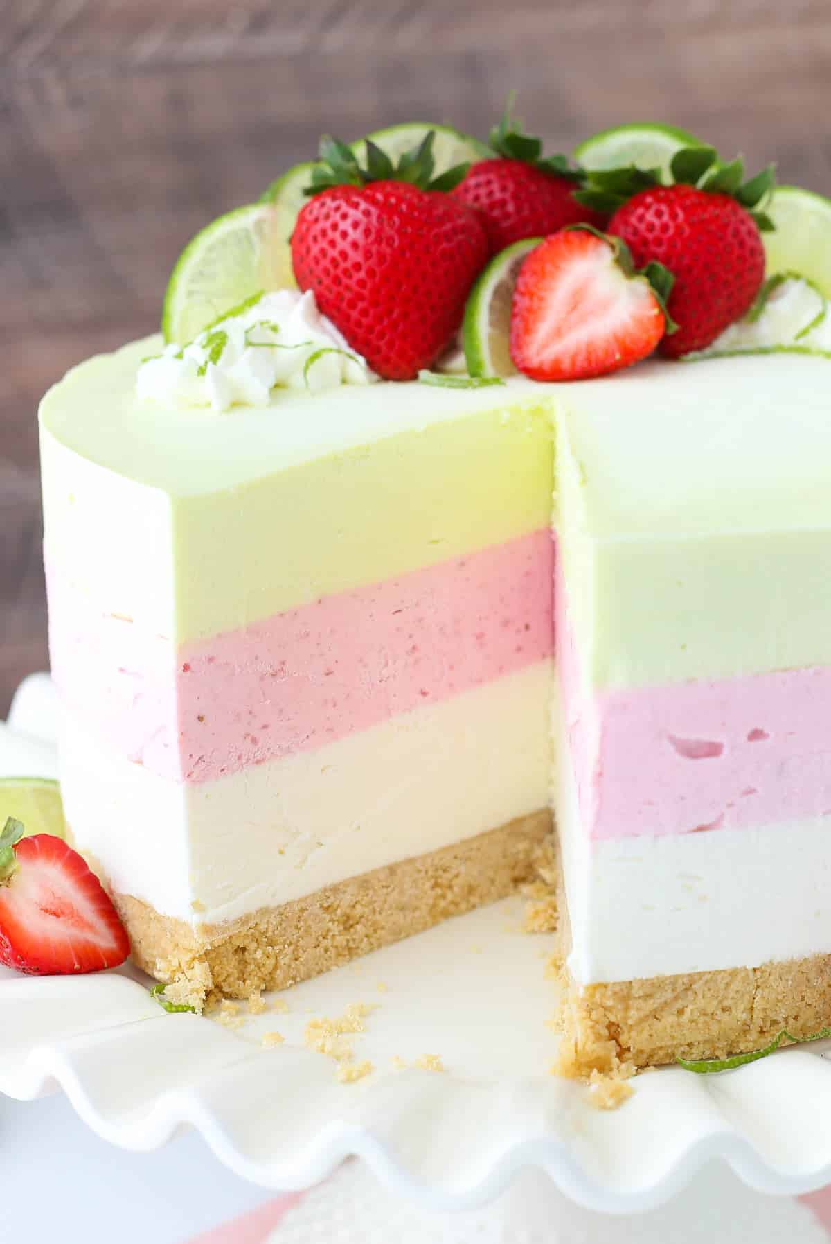 This Key Lime Strawberry Coconut Ice Cream Cake Recipe combines three delicious flavors into one colorful no-bake, no-churn treat! A key lime strawberry coconut ice cream cake on a white cake stand with a slice removed to show layers.
