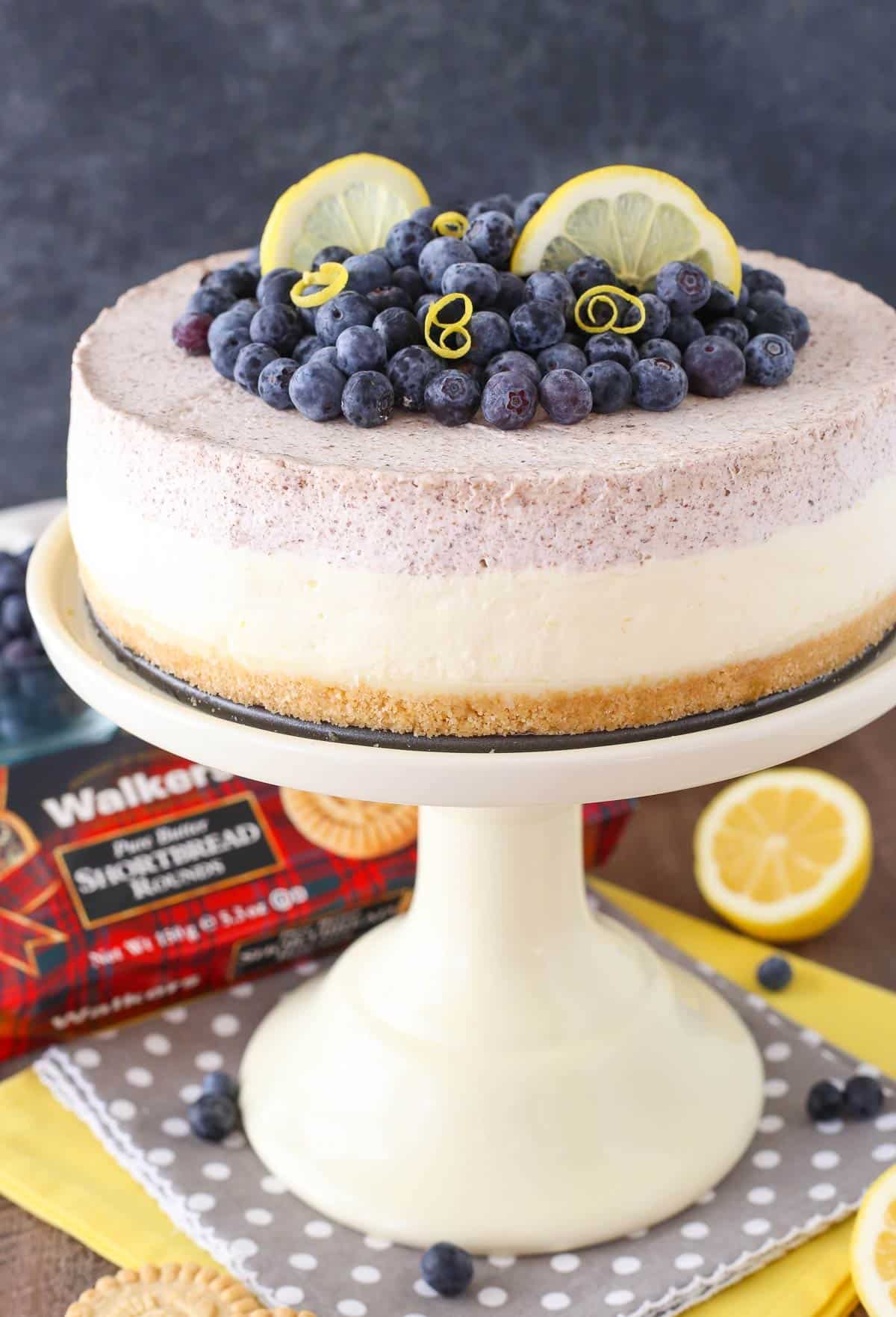 Side view of a lemon blueberry shortbread mousse cake on a white cake stand next to a box of shortbread.