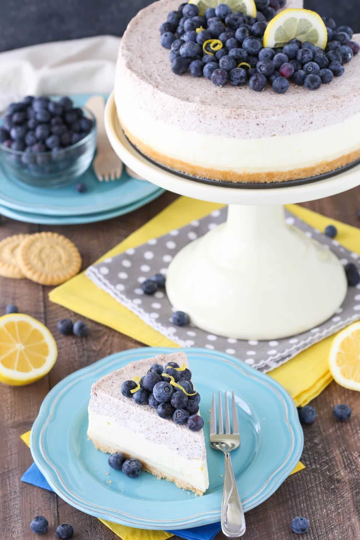 A slice of lemon blueberry shortbread mousse cake on a blue plate with the rest of the cake in the background.