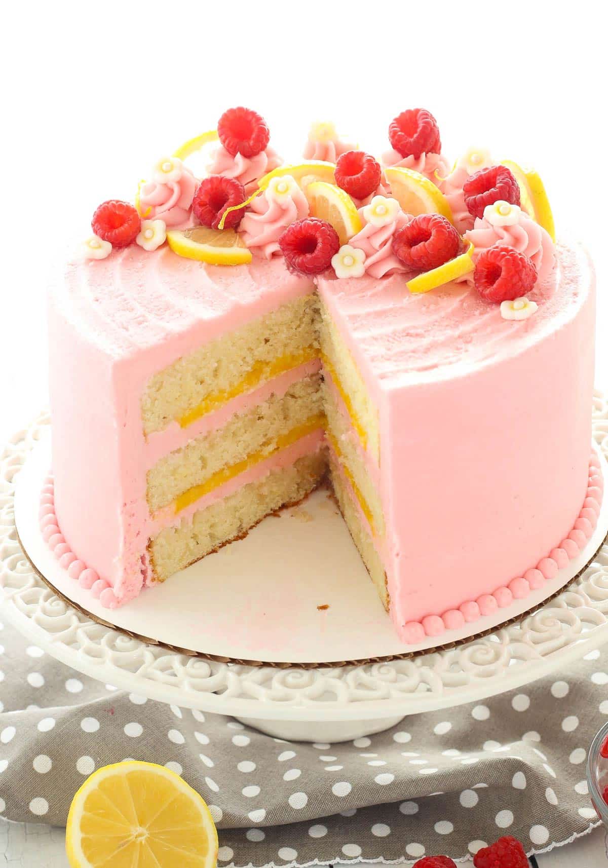 A lemon raspberry cake on a white cake stand with a slice removed to show interior layers.
