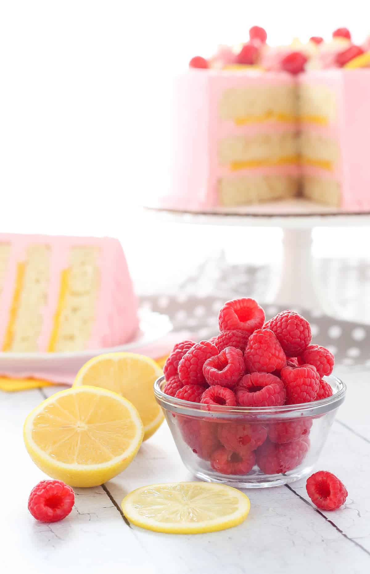 A bowl of raspberries next to sliced lemons with a lemon raspberry cake in the background.