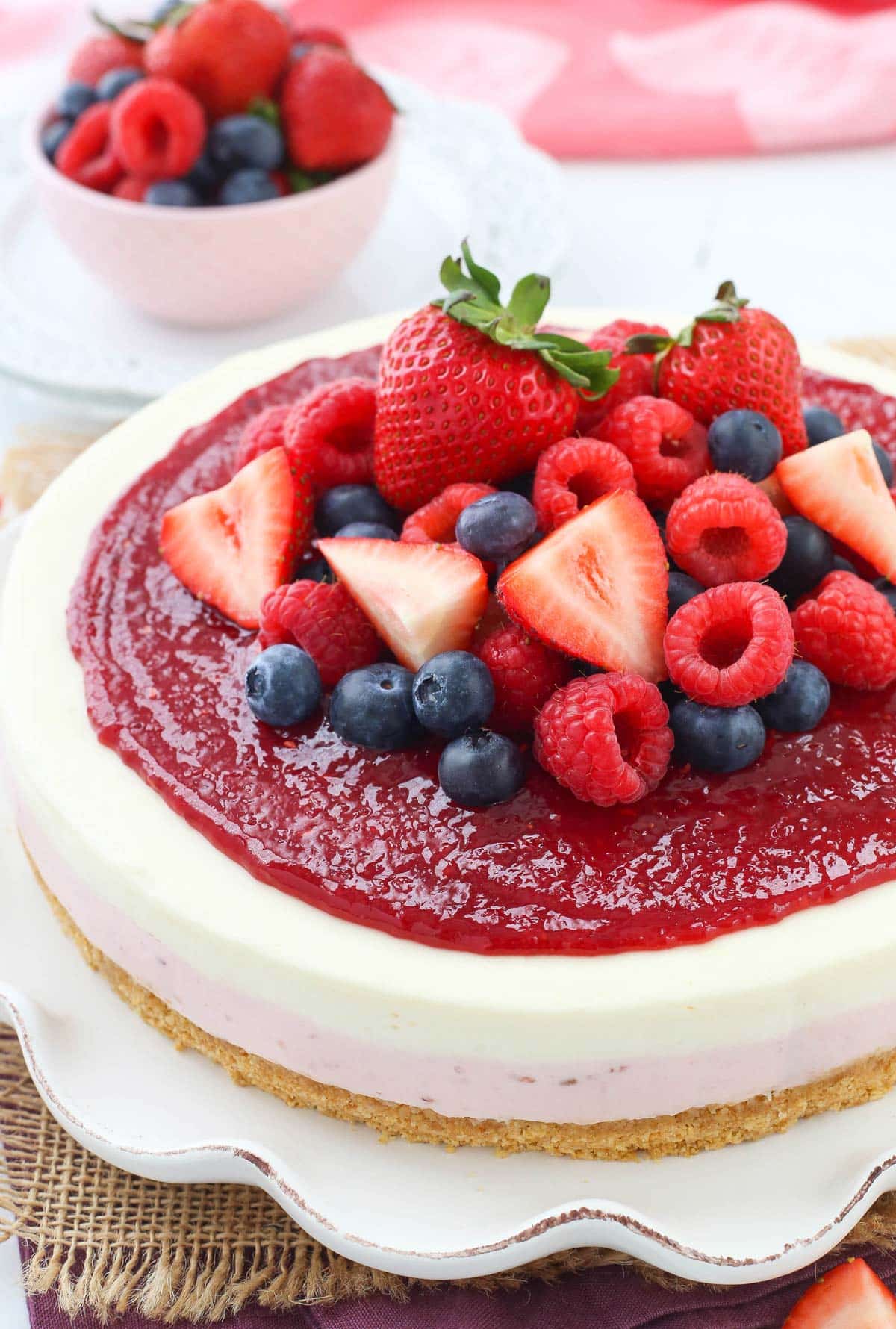 A no bake berry vanilla cheesecake on a white ruffled cake plate.