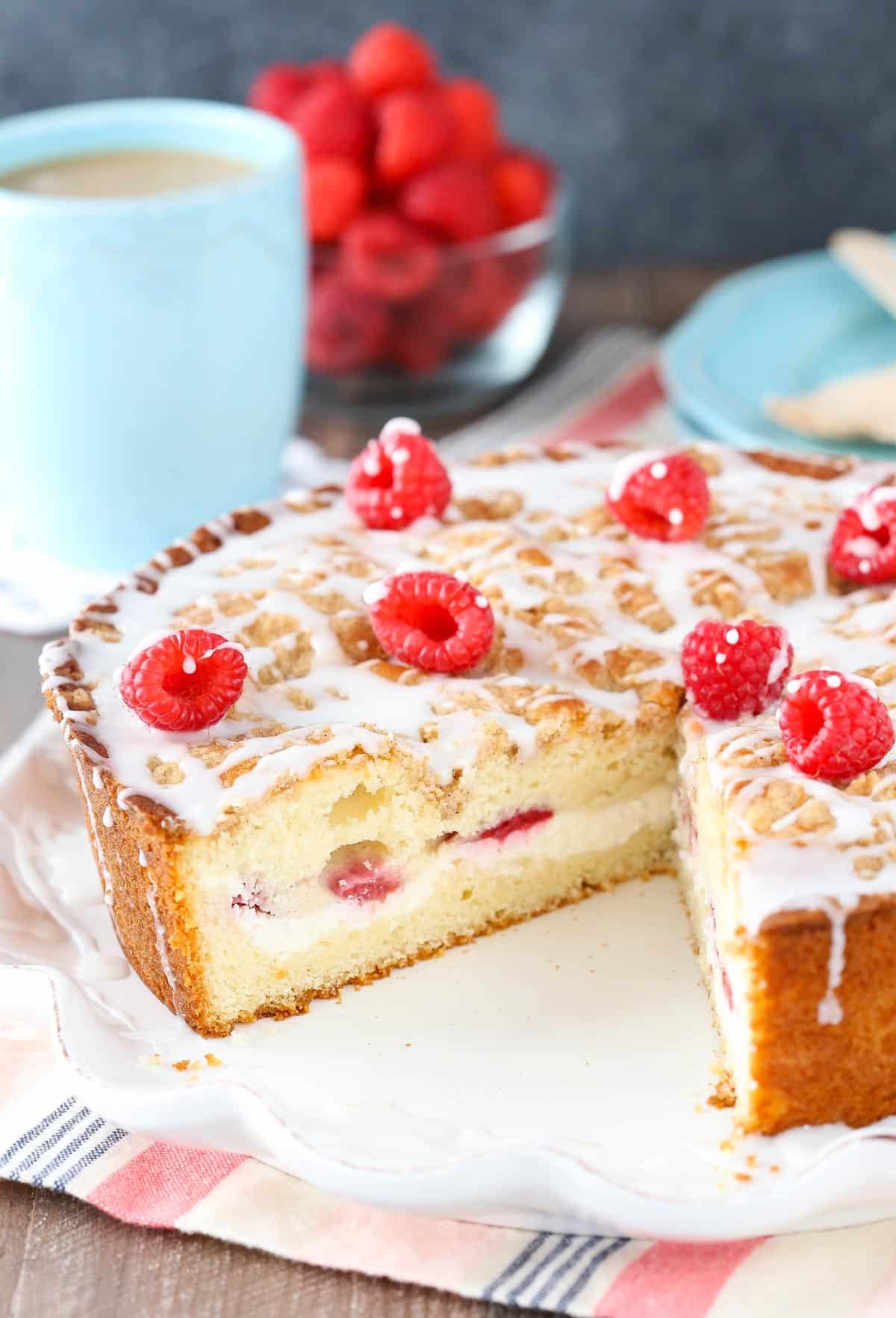 A raspberry cream cheese coffee cake on a white ruffled cake plate with a slice removed to show internal layers.