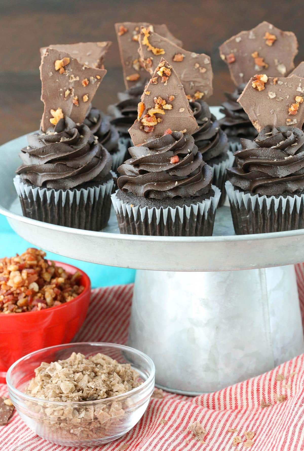 A silver cake plate covered with smokey bourbon chocolate cupcakes with bacon bark.