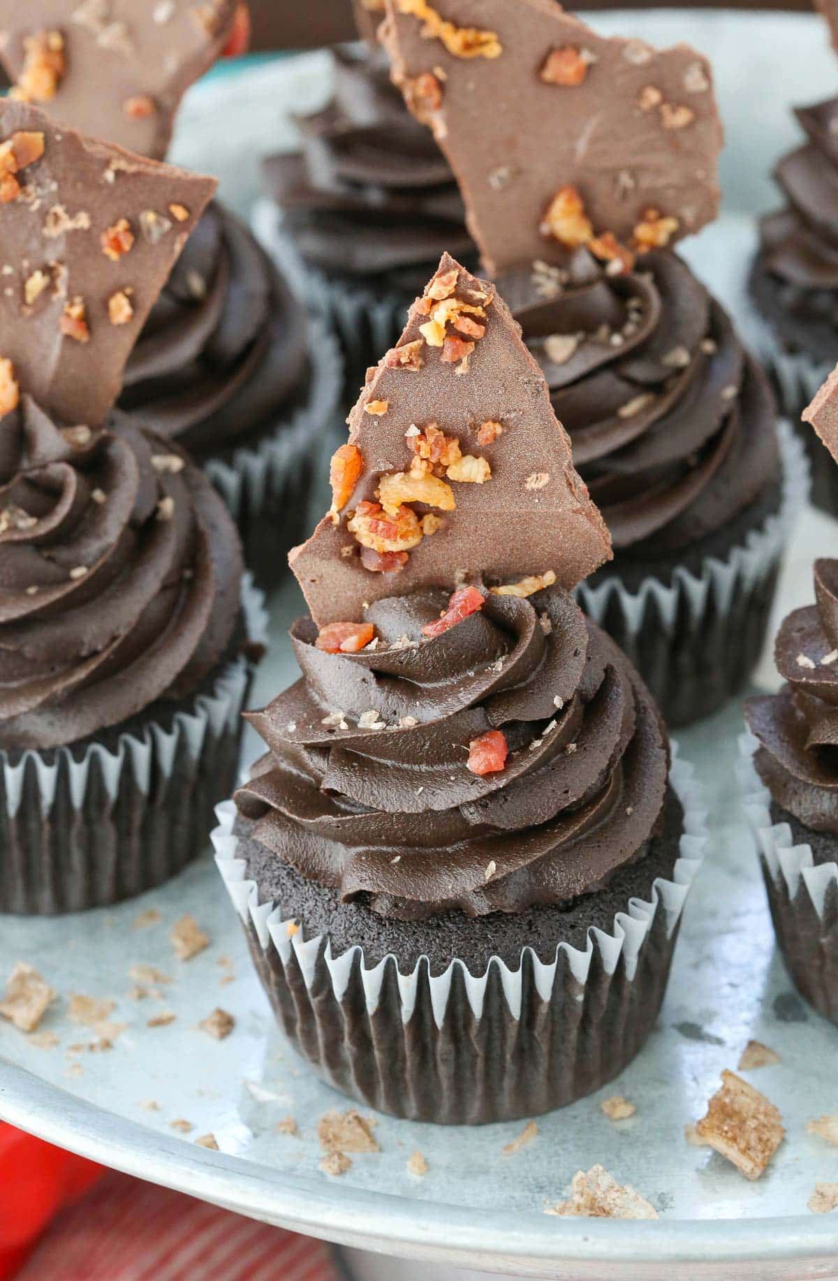 Several smokey bourbon chocolate cupcakes with bacon bark on a silver cake plate.