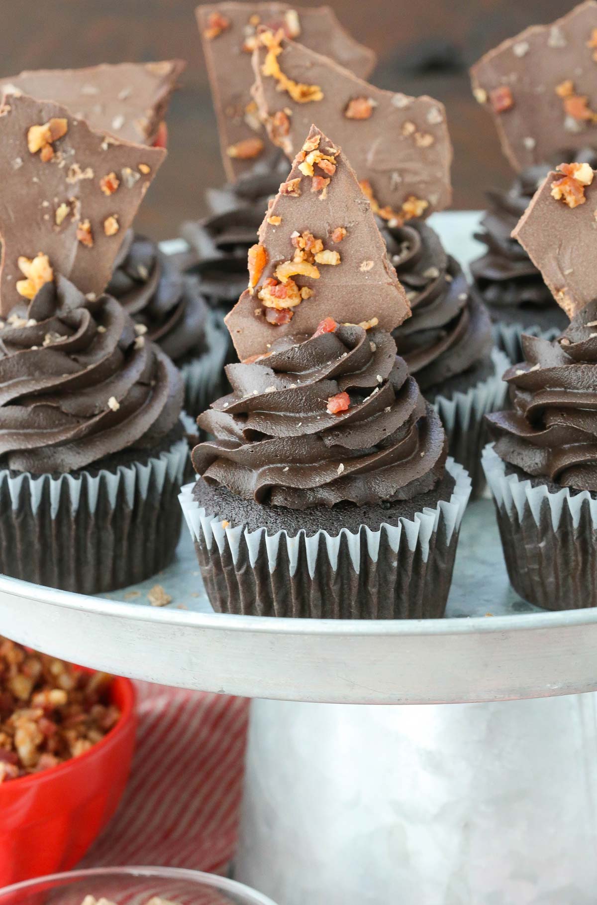 Close up of smokey bourbon chocolate cupcakes with bacon bark on a silver cake stand.
