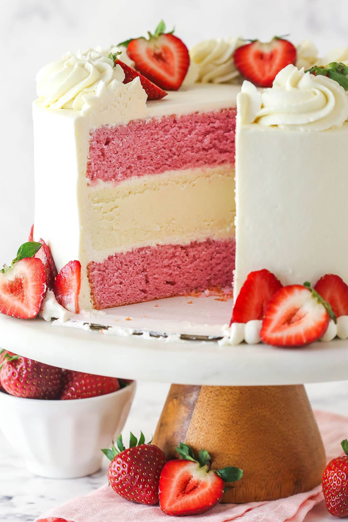This strawberries and cream cheesecake cake layers strawberry cake, vanilla cheesecake, and cream cheese frosting for one incredible dessert. Strawberries and cream cheesecake cake on a cake stand with a slice taken out of it.