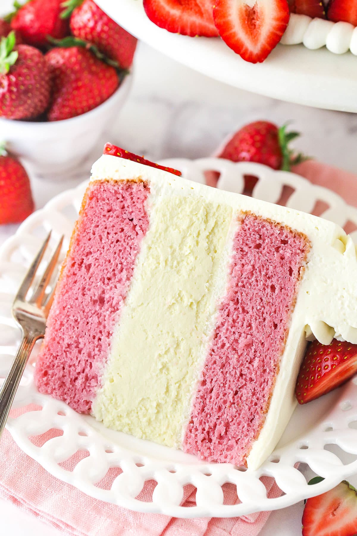 This strawberries and cream cheesecake cake layers strawberry cake, vanilla cheesecake, and cream cheese frosting for one incredible dessert. A slice of strawberries and cream cheesecake cake on a plate.