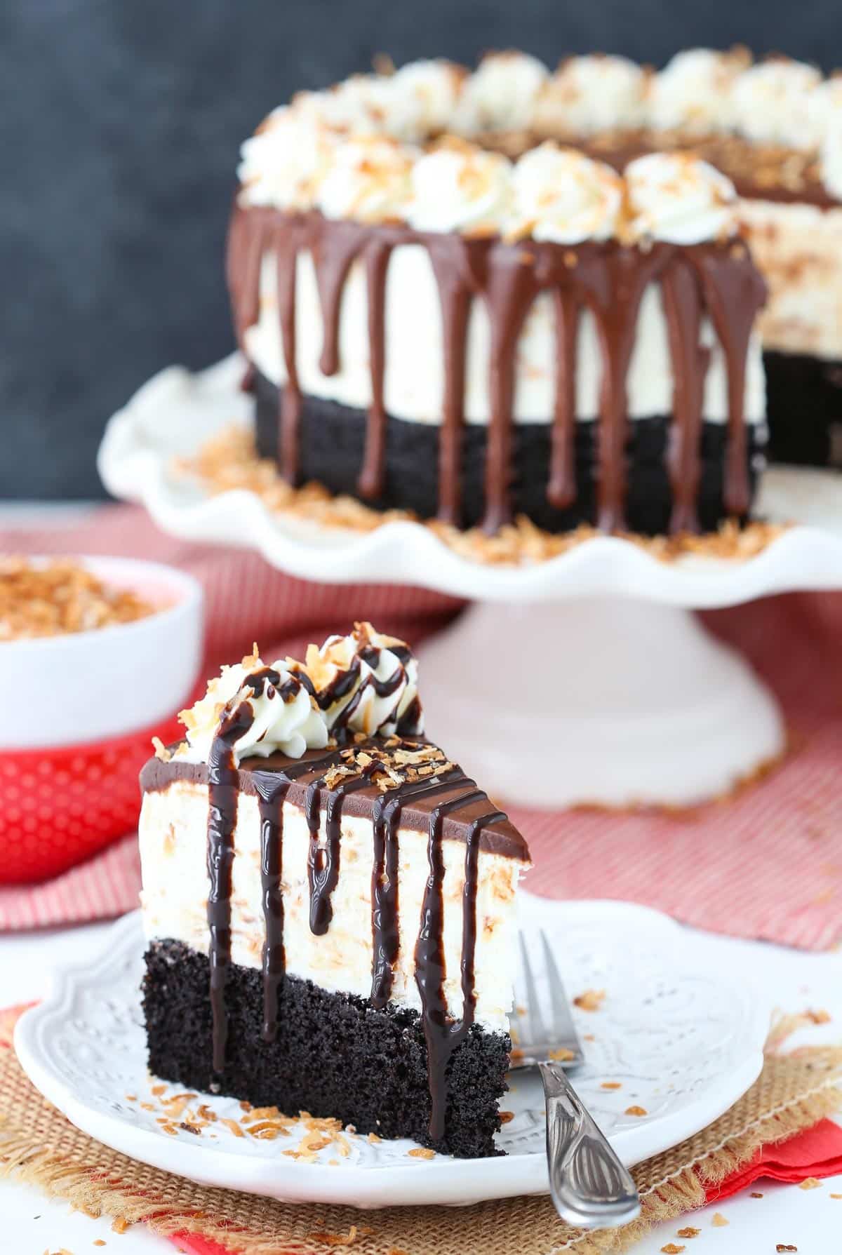 A slice of toasted coconut chocolate ice cream cake on a white plate with the rest of the cake in the background.