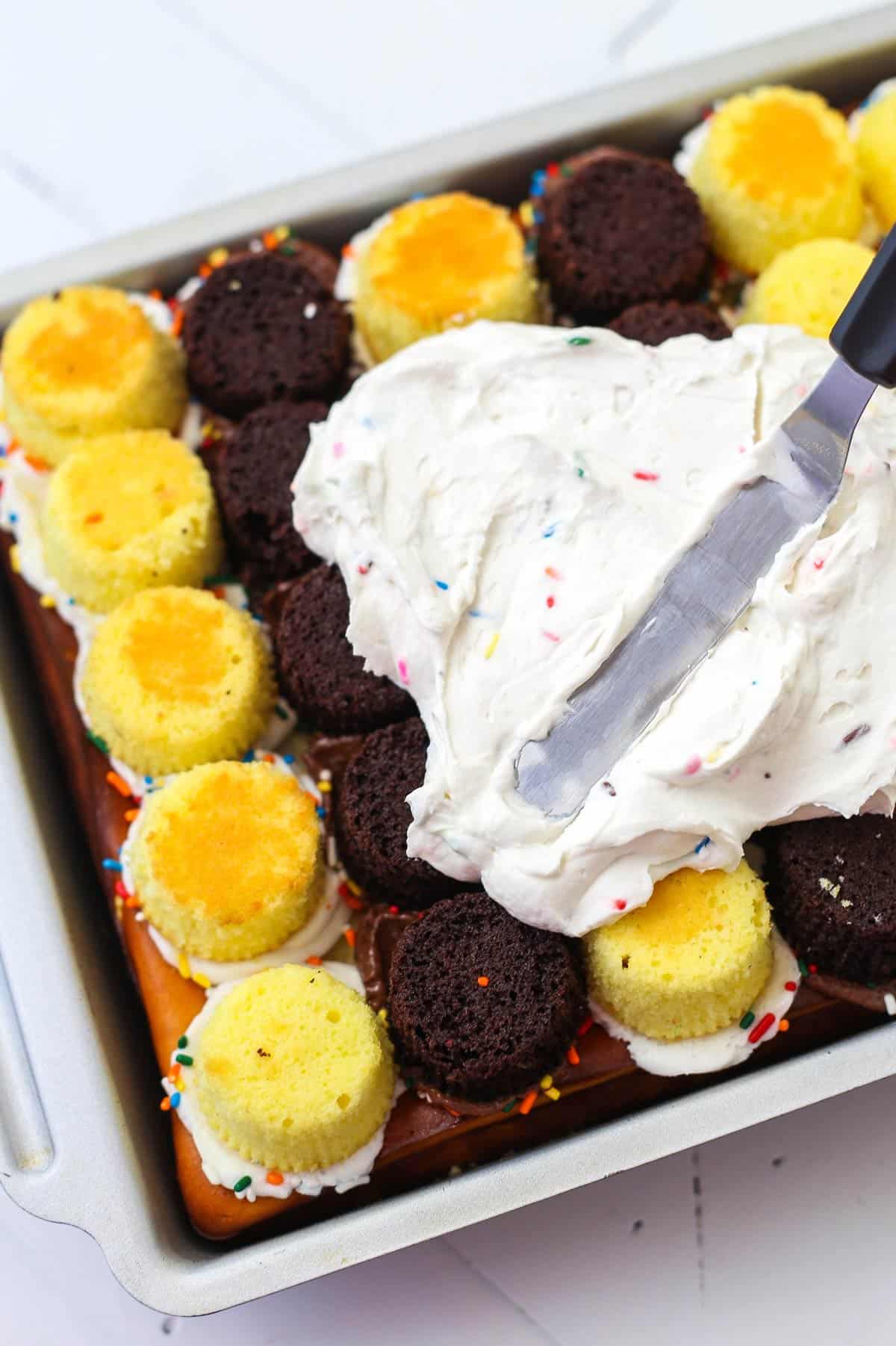 This epic birthday cake combines chewy chocolate chip cookies, creamy Funfetti cheesecake, moist cupcakes & fluffy cake batter whipped cream! A spatula spreading frosting on top of an ultimate birthday cake.