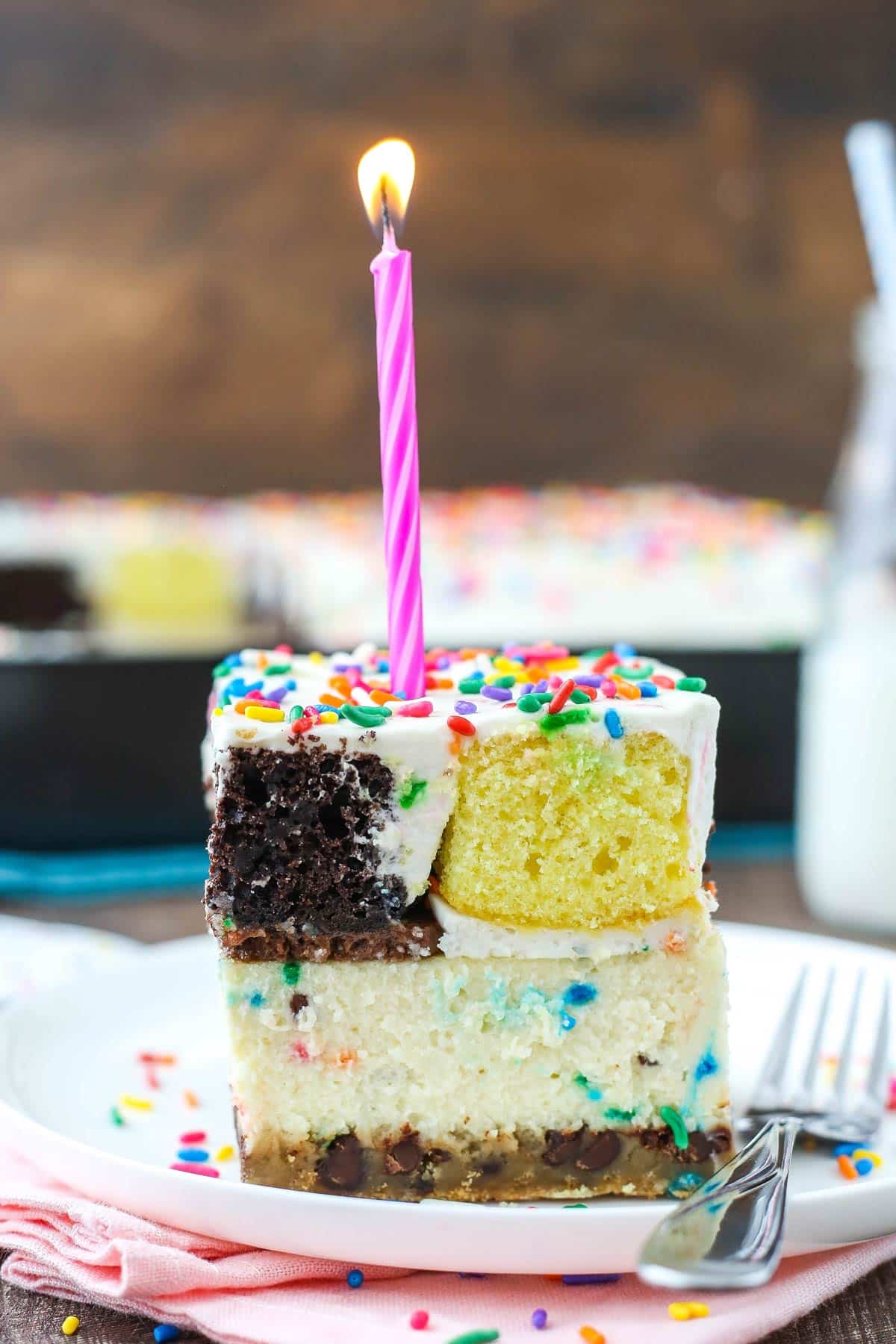 This epic birthday cake combines chewy chocolate chip cookies, creamy Funfetti cheesecake, moist cupcakes & fluffy cake batter whipped cream! A slice of ultimate birthday cake with a candle on top.