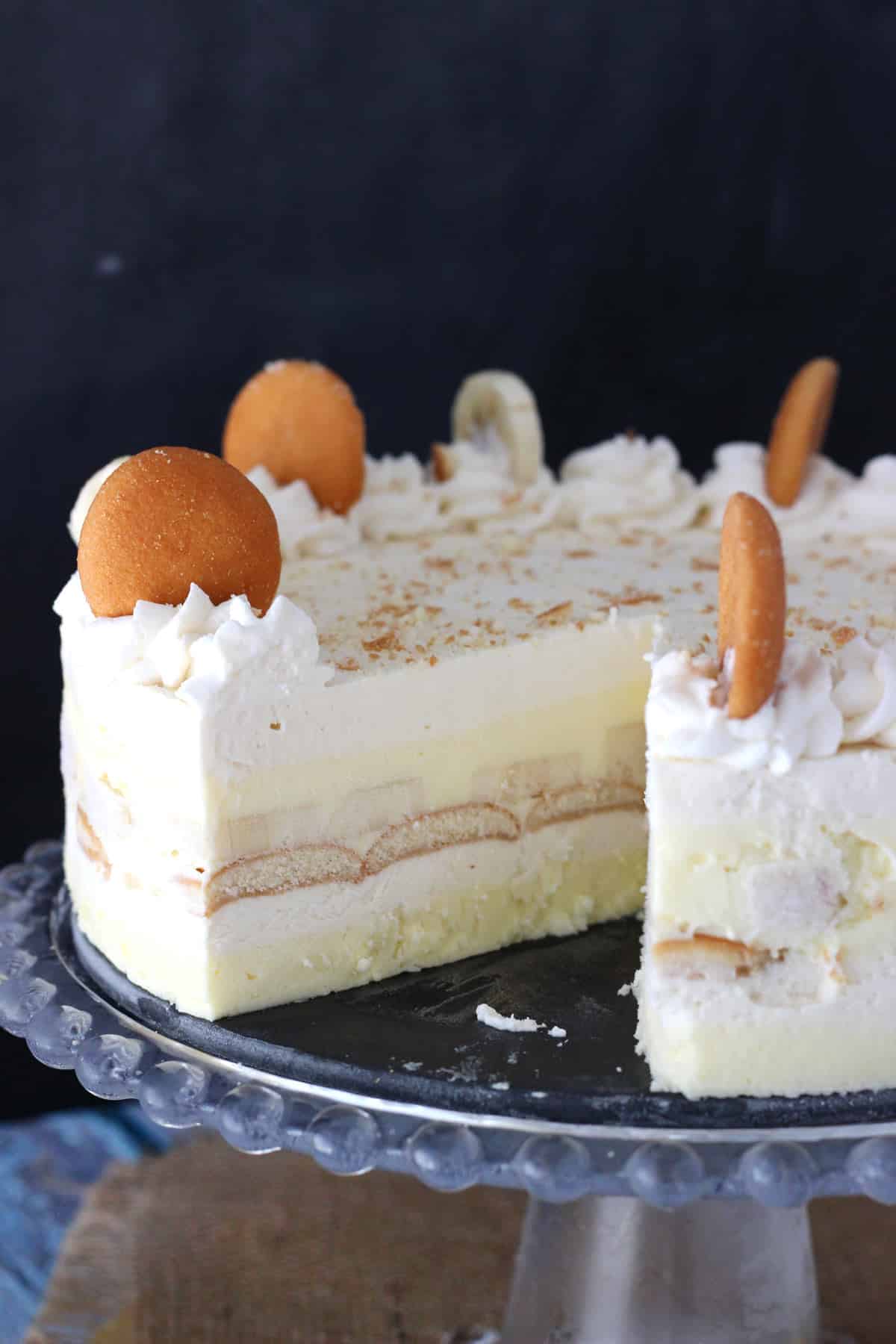 A banana pudding icebox cake on a glass cake stand with a slice removed to show layers.