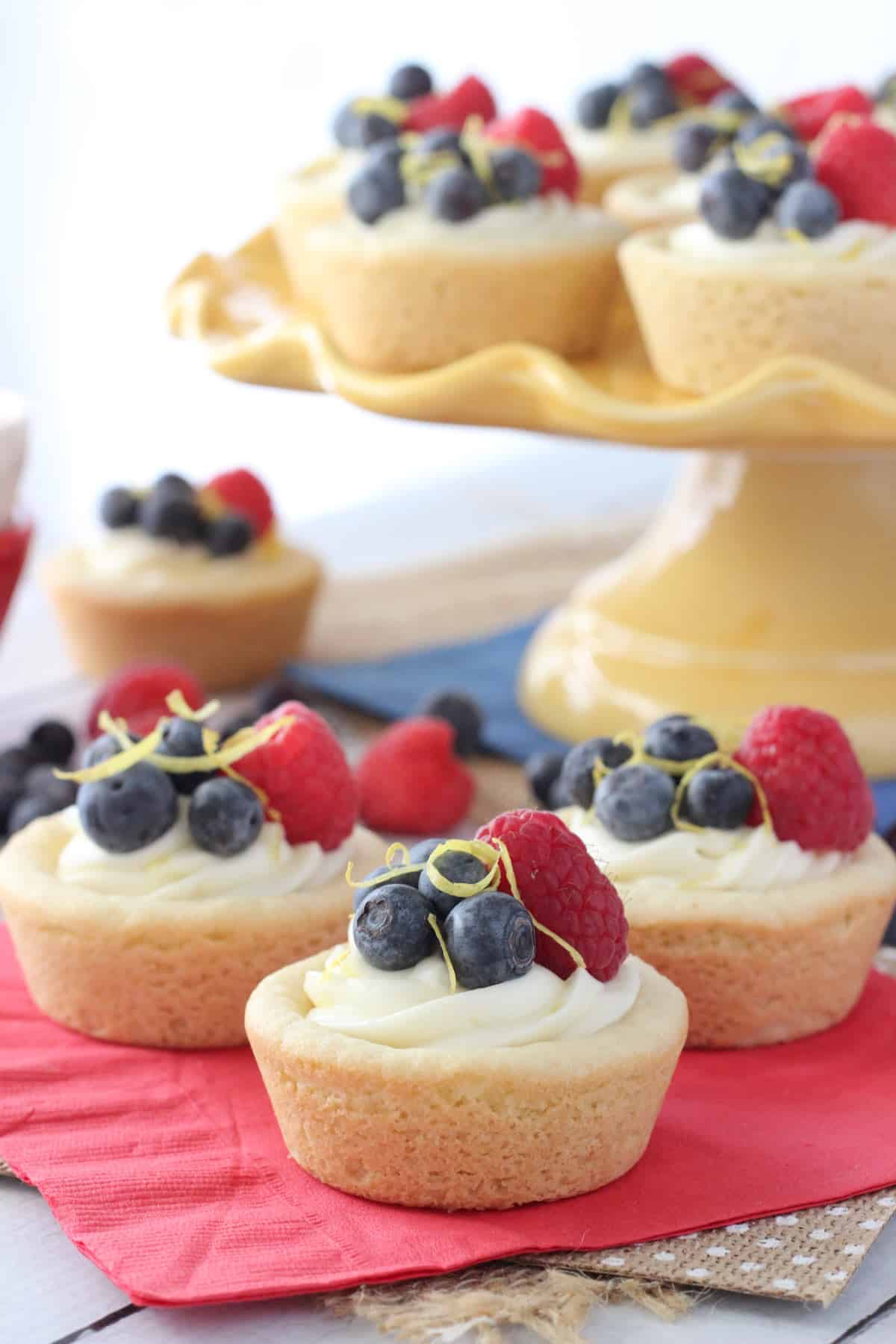 Three berry lemon cheesecake cookie cups on a red napkin with the rest of the cups on a yellow cake stand.