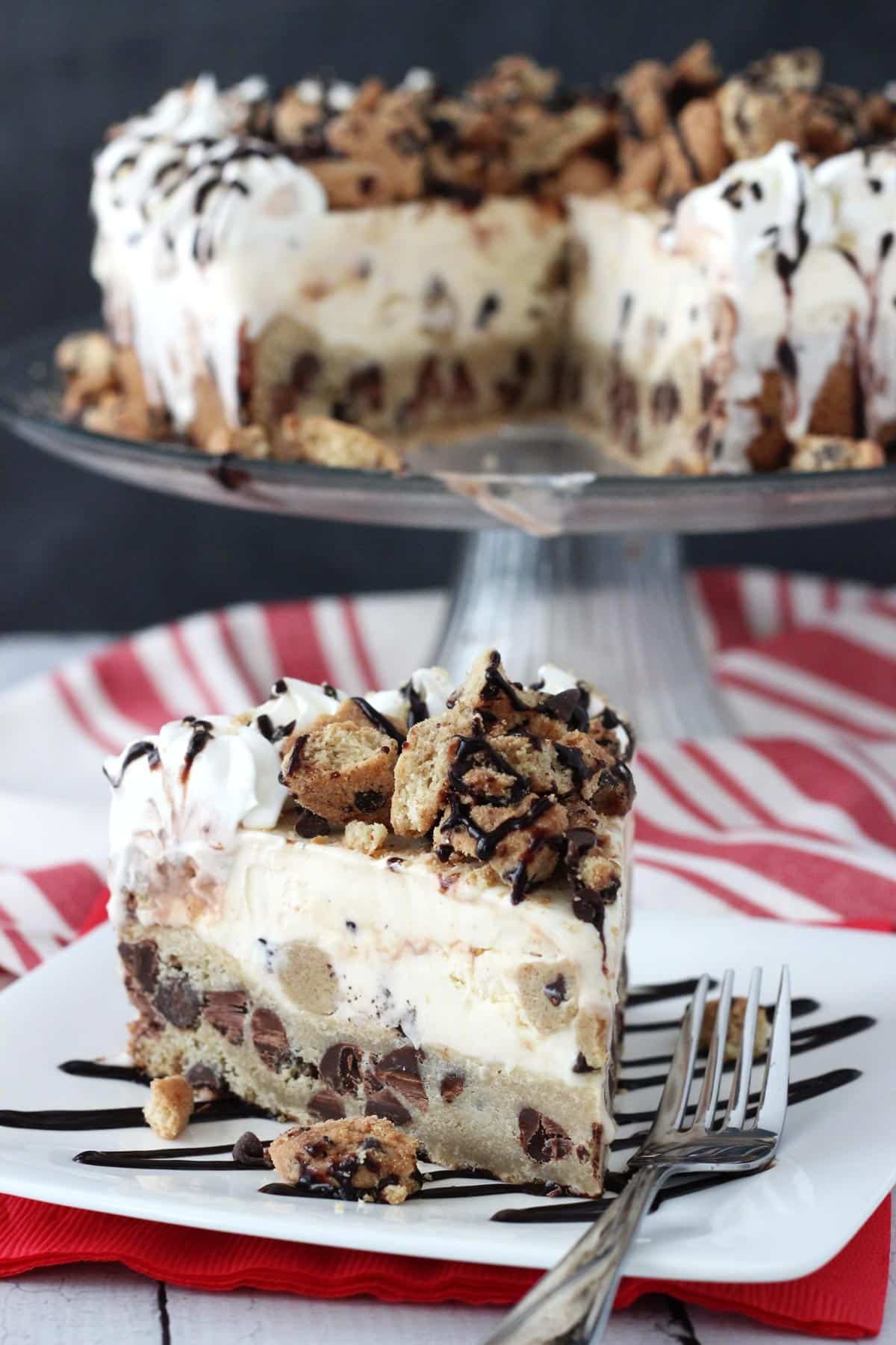 A slice of chocolate chip cookie ice cream cake on a white plate with the rest of the cake in the background.