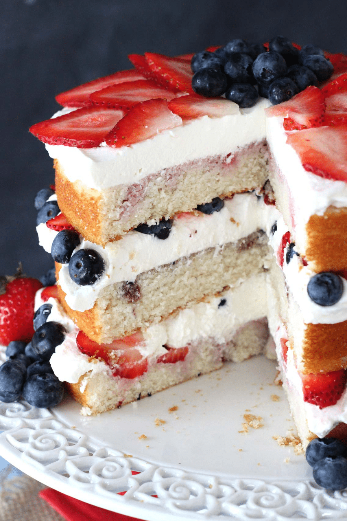 A fresh berry vanilla layered cake on a white cake stand with a few slices removed to show internal layers.