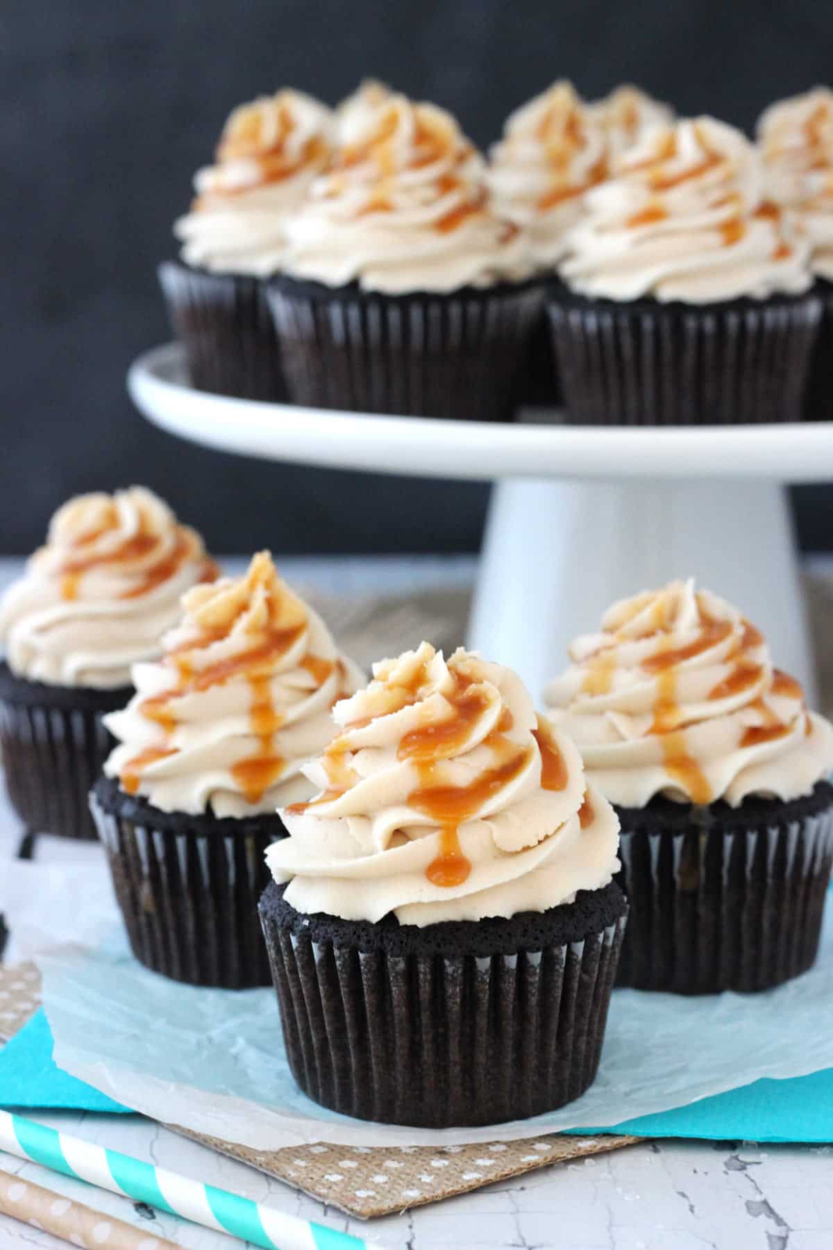Four Kahlua chocolate cupcakes with salted caramel with a cake plate of more cupcakes in the background.