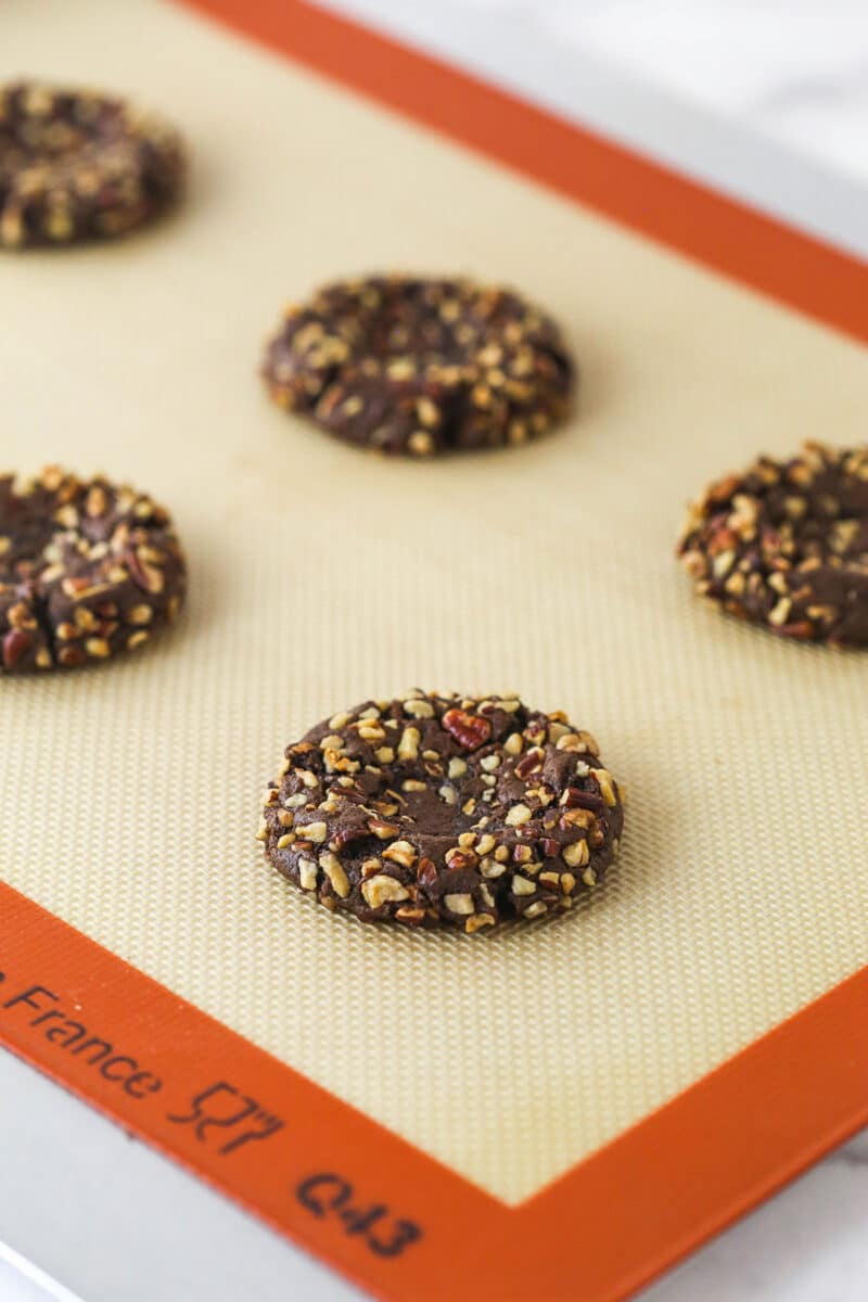 These decadent turtle thumbprint cookies feature a rich chocolate base, gooey caramel center, and crunchy pecan coating. Wow! chocolate pecan crusted cookie on a silicone mat with a divot in the center of each.