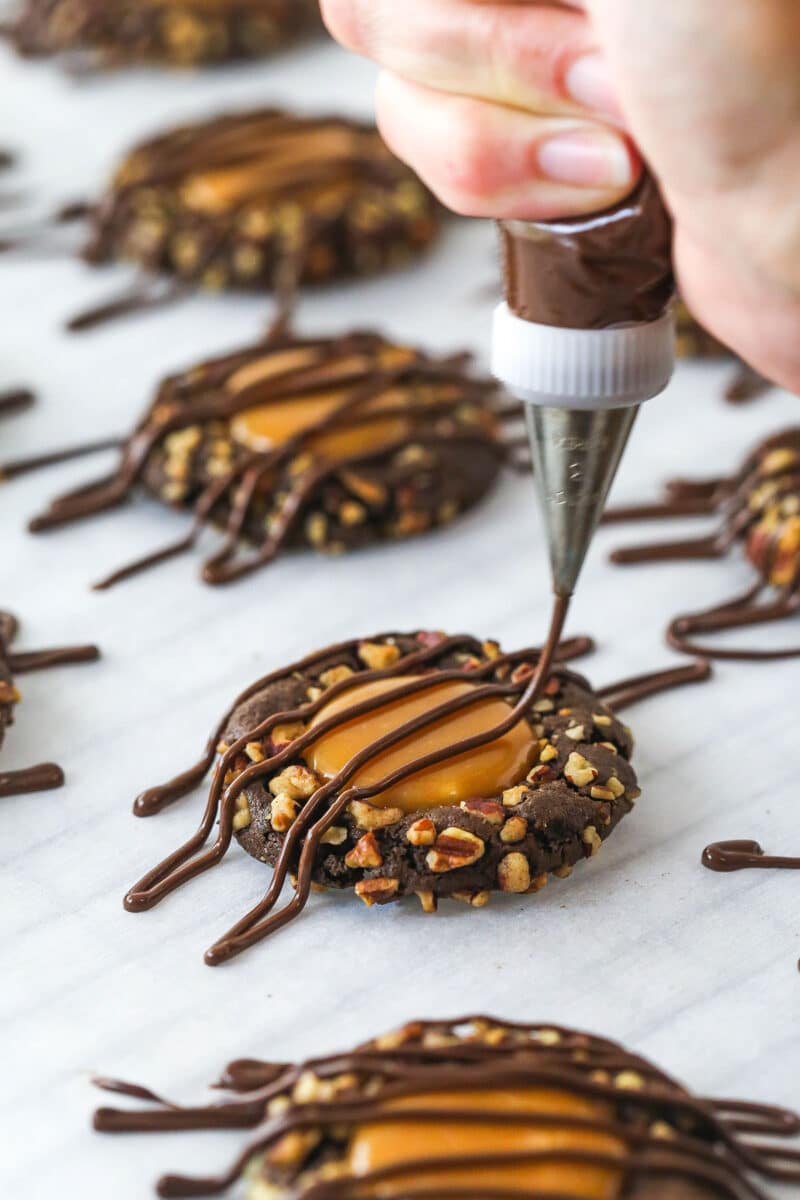 These decadent turtle thumbprint cookies feature a rich chocolate base, gooey caramel center, and crunchy pecan coating. Wow! Piping chocolate ganache over turtle thumbprint cookies.