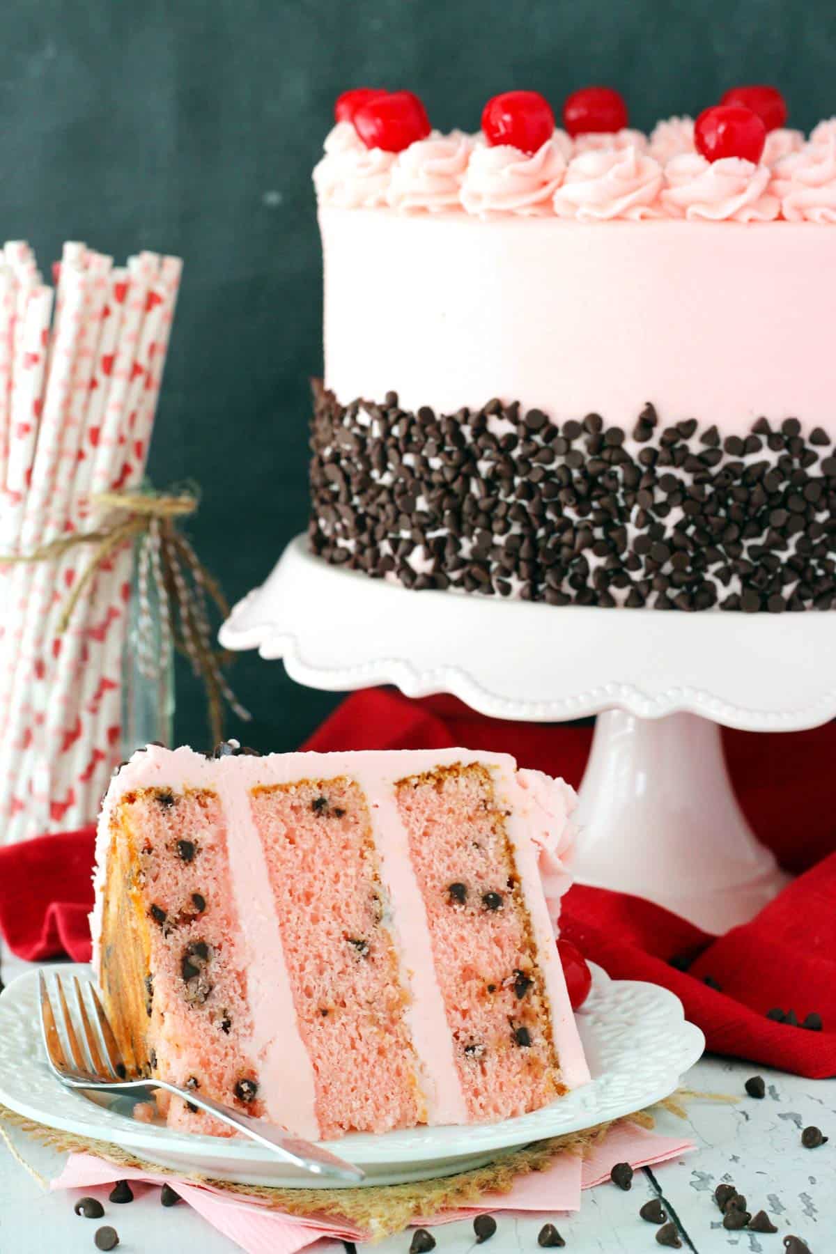 A slice of cherry chocolate chip cake on a small plate with the rest of the cake in the background.