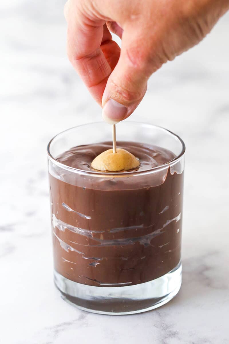These Buckeyes have the perfect smooth and creamy texture with a sweet, nutty peanut butter and chocolate flavor. No baking required! A hand using a toothpick to dip a peanut butter ball into melted chocolate.