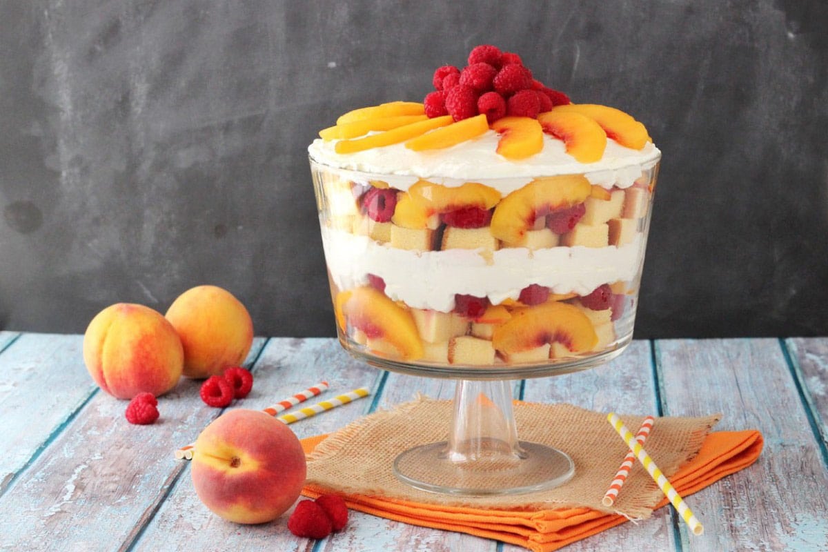 Raspberry Peach Sangria Trifle A raspberry peach sangria trifle in a glass trifle bowl next to peaches and raspberries.