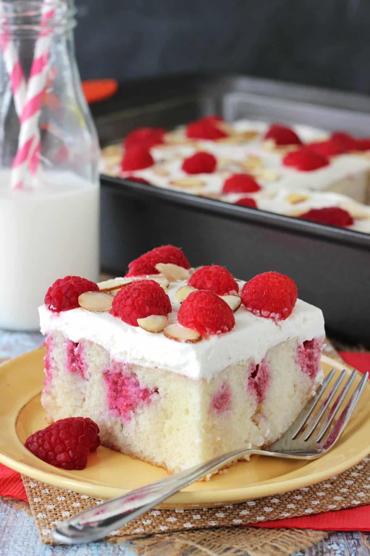 A slice of raspberry almond poke cake on a yellow plate with the rest of the cake in the background.