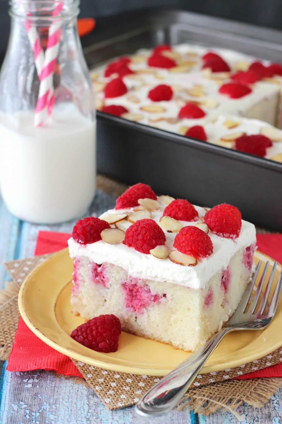 A slice of raspberry almond poke cake on a yellow plate with the rest of the cake in the background.