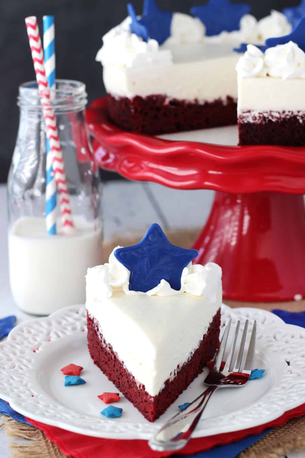 A slice of red velvet blondie cheesecake on a small white plate with the rest of the cake in the background.