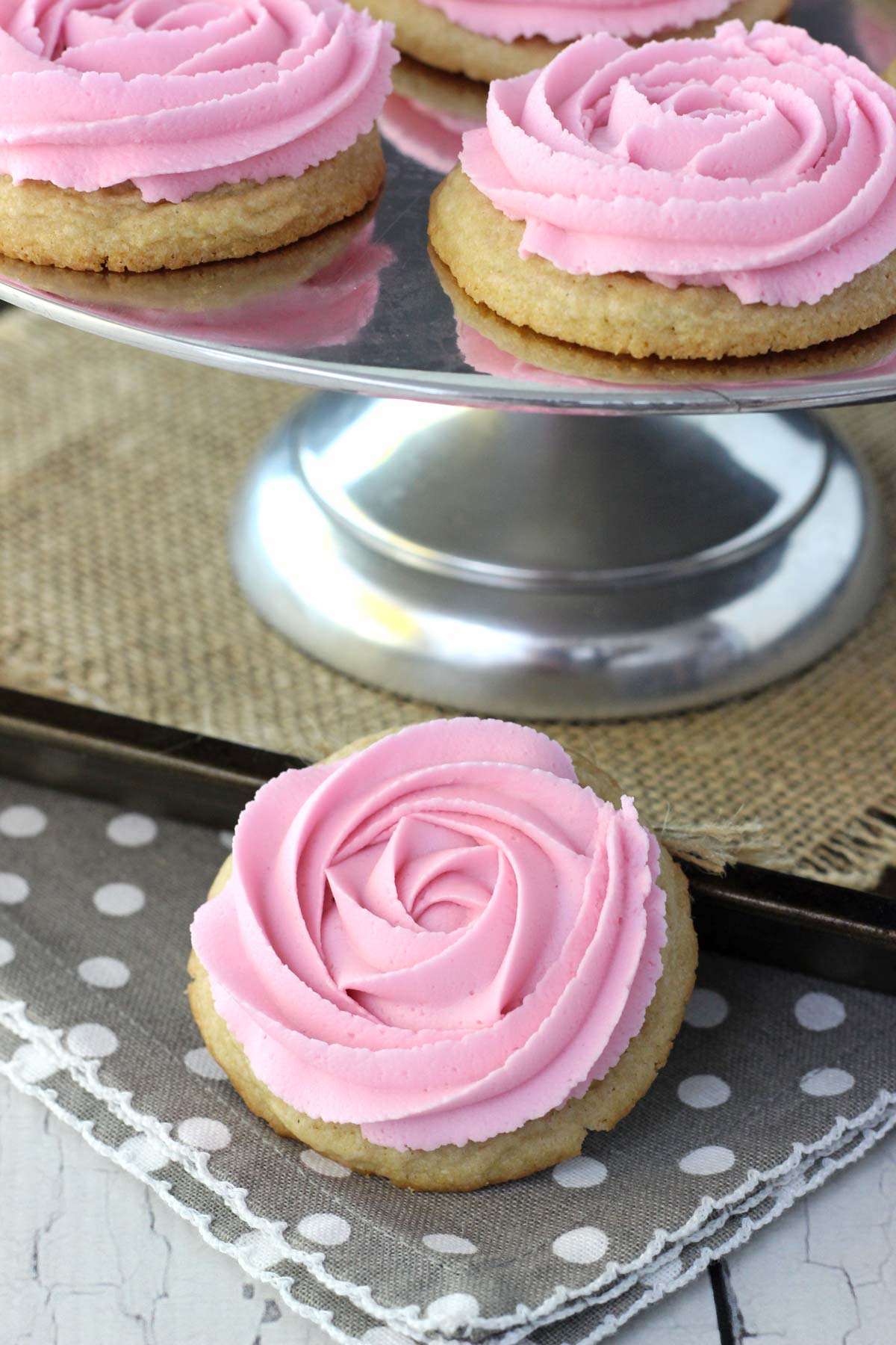 Close up of a rosette sugar cookie below a silver cake stand topped with more cookies.