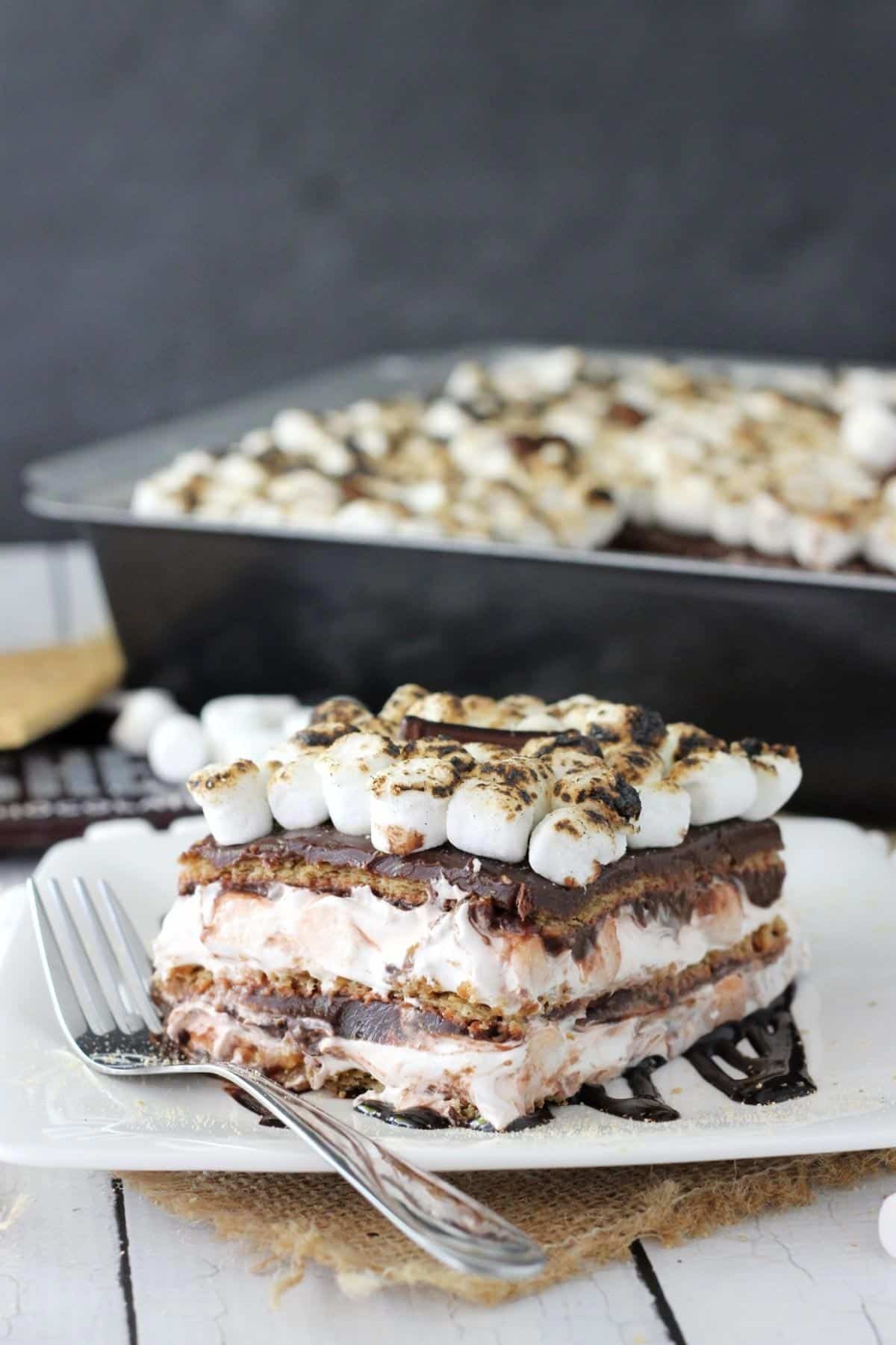 Close up of a slice of smores icebox cake on a white plate with the rest of the cake in the background.
