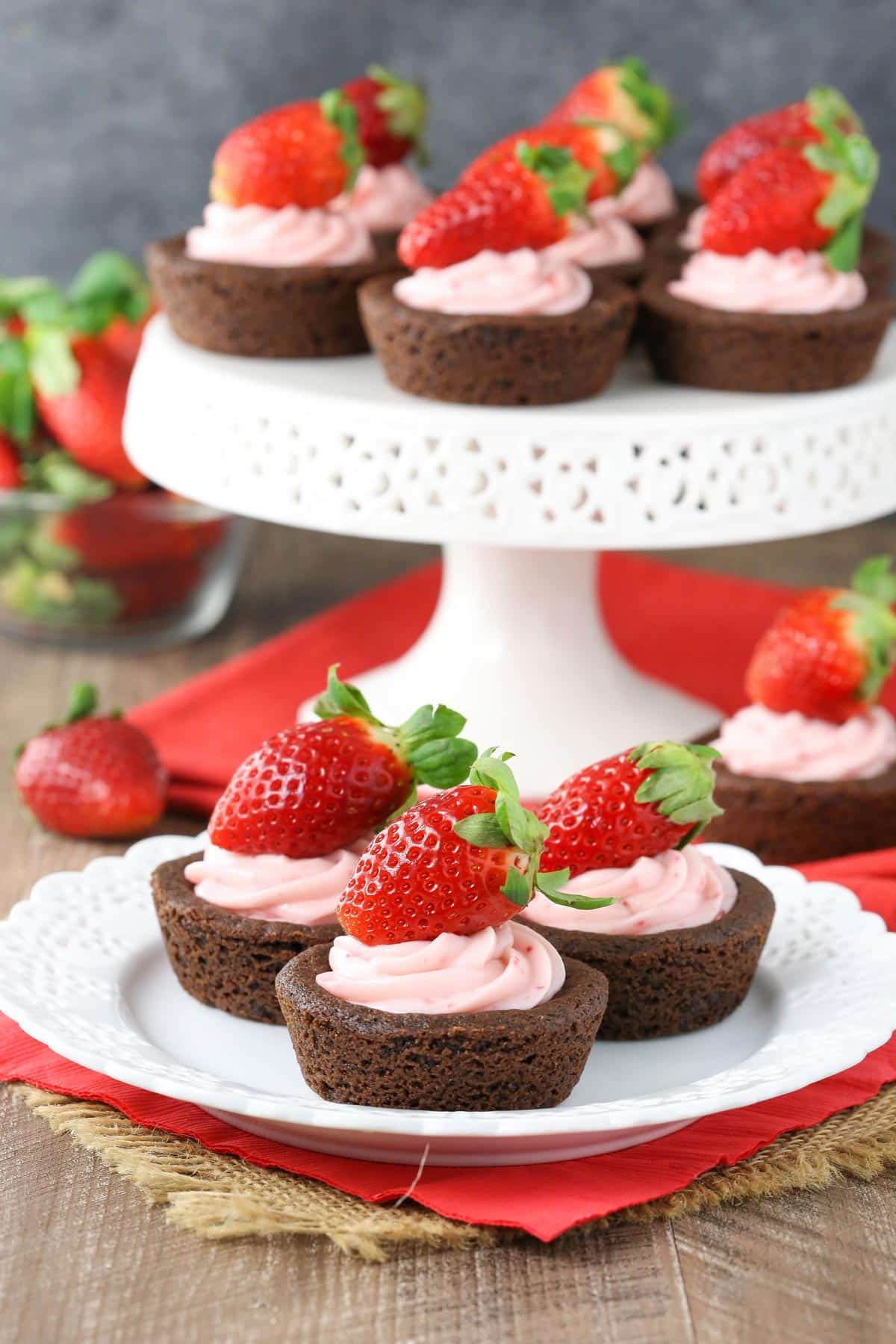 A few strawberry cheesecake chocolate cookie cups on a small white plate with more in the background on a white cake stand.
