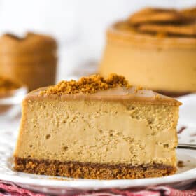 A slice of Biscoff cheesecake on a white plate in front of the full cheesecake