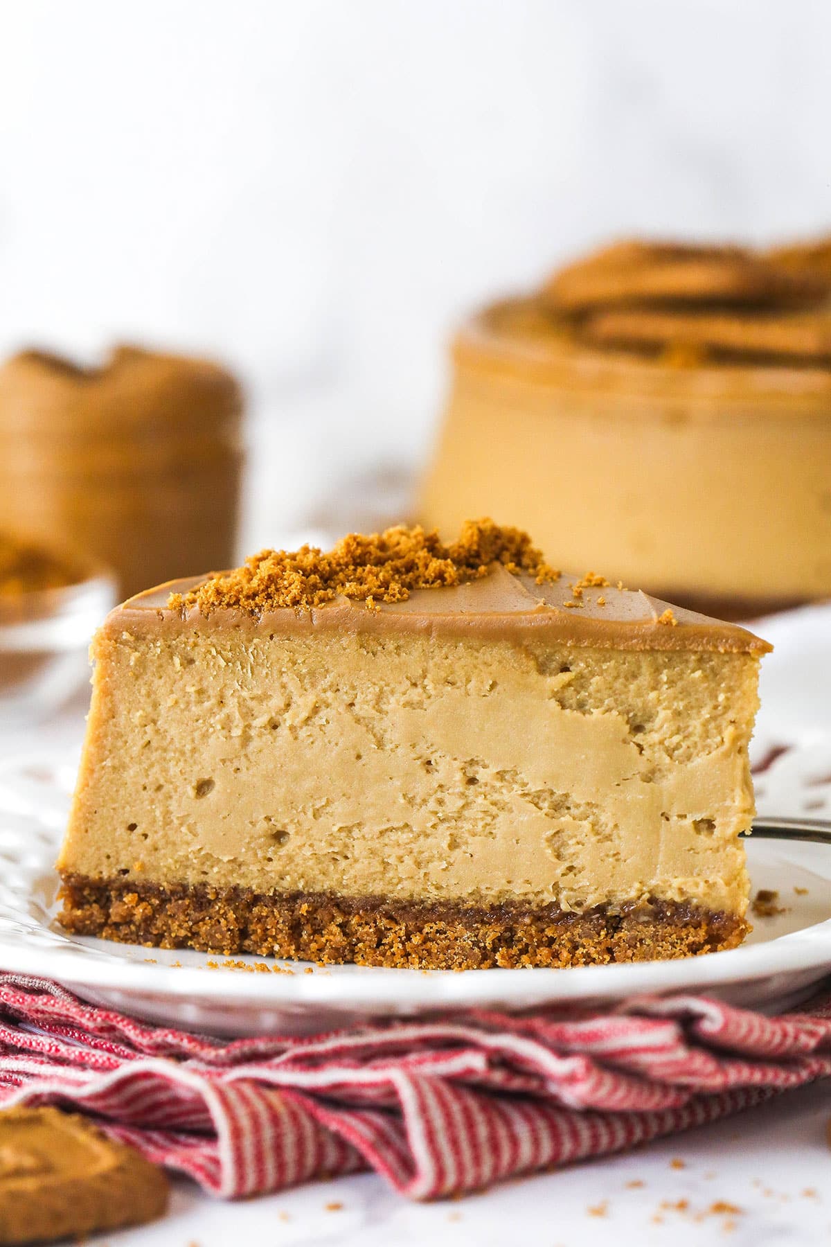 With a Biscoff cookie crust and cookie spread in the filling, this Biscoff Cheesecake tastes just like the cookies. A slice of Biscoff cheesecake on a white plate in front of the full cheesecake