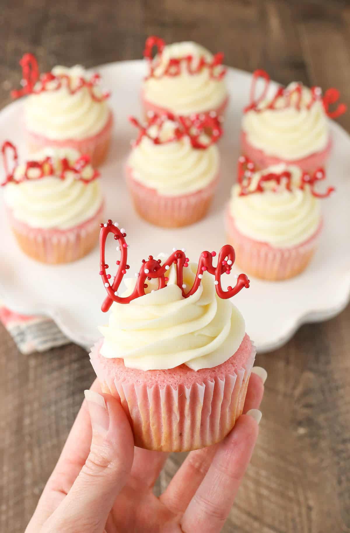 Hand holding up a strawberry cupcake with cream cheese frosting with more cupcakes in the background.