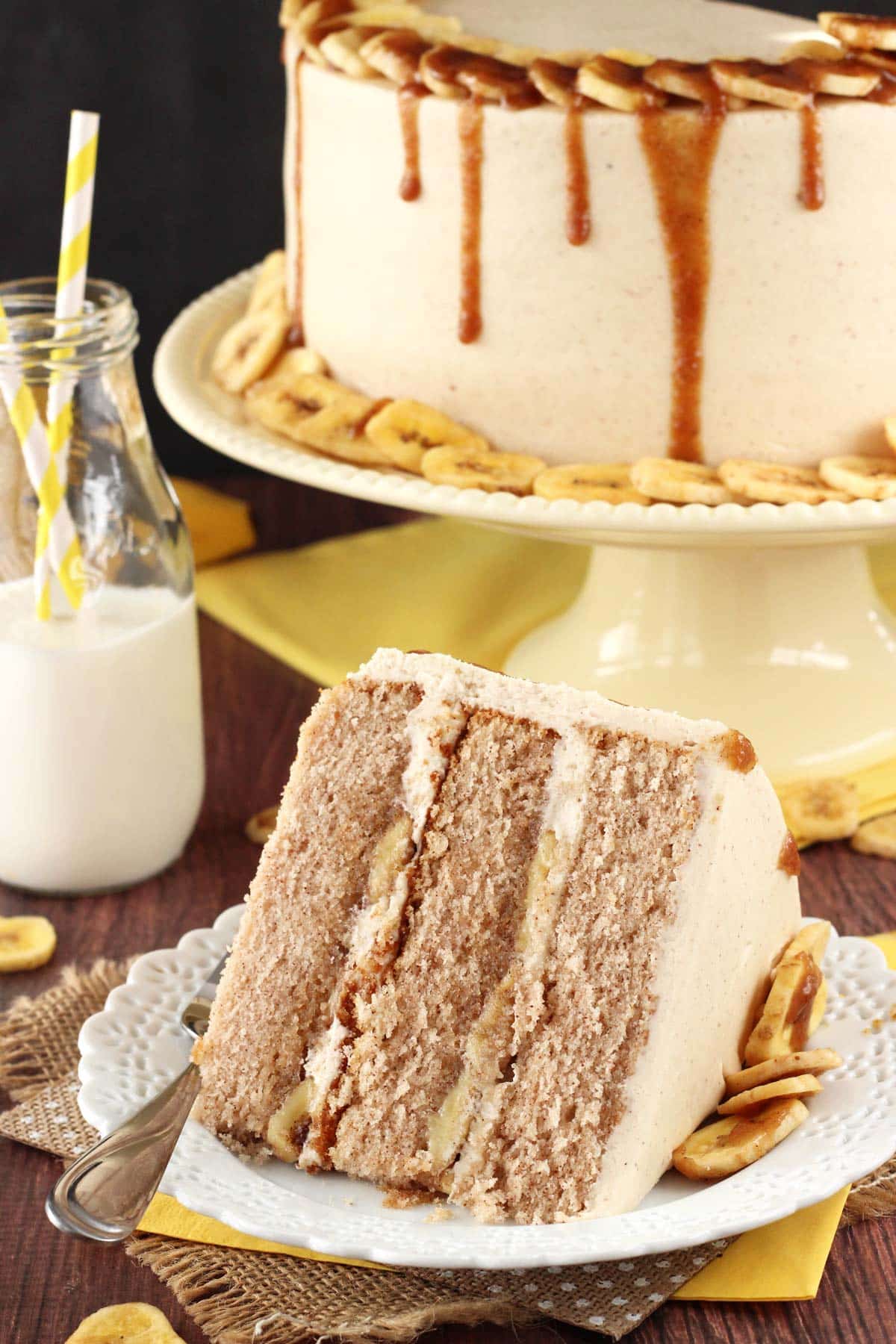 A slice of bananas foster layer cake on a small white plate with the rest of the cake in the background on a cream colored cake stand.