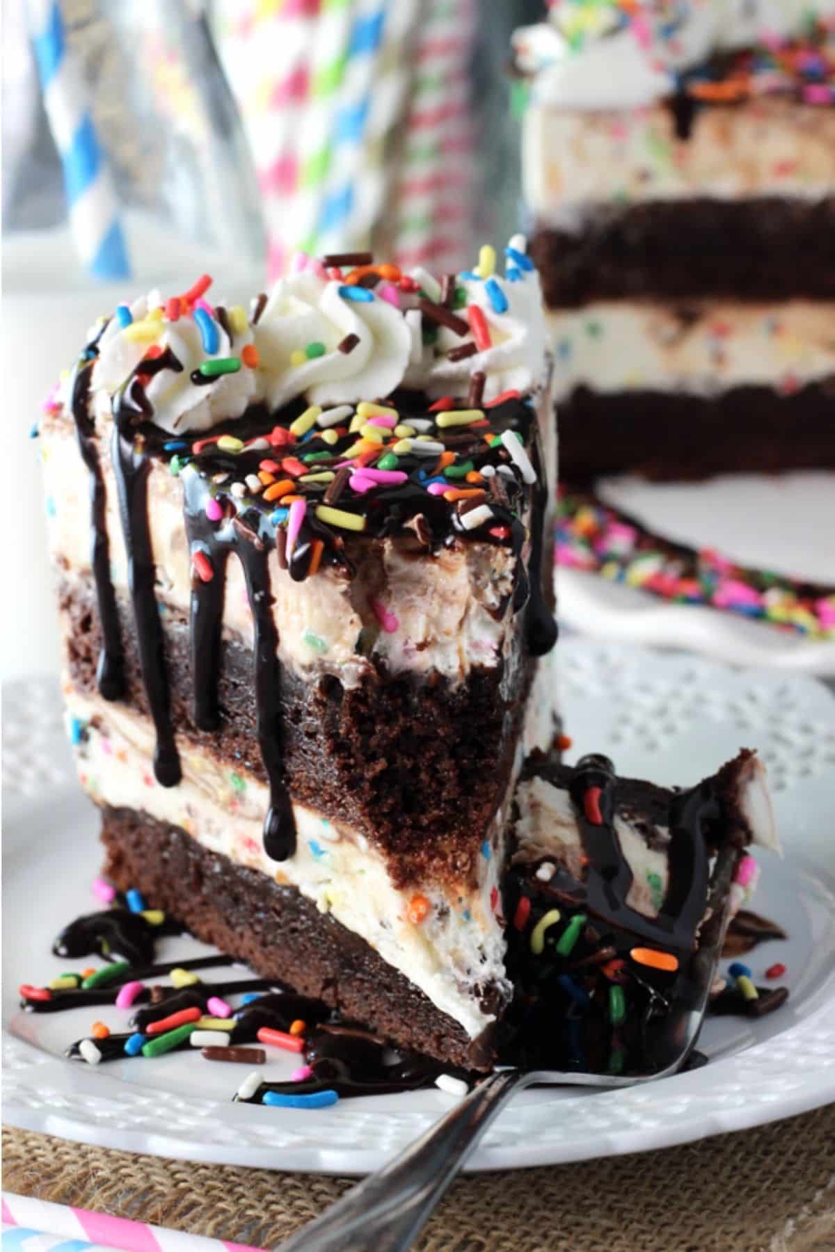 This Cake Batter Fudge Brownie Ice Cream Cake combines funfetti cake batter, brownies, fudge sauce and ice cream into one dessert! A slice of cake batter fudge brownie ice cream cake with a bite removed on a fork.