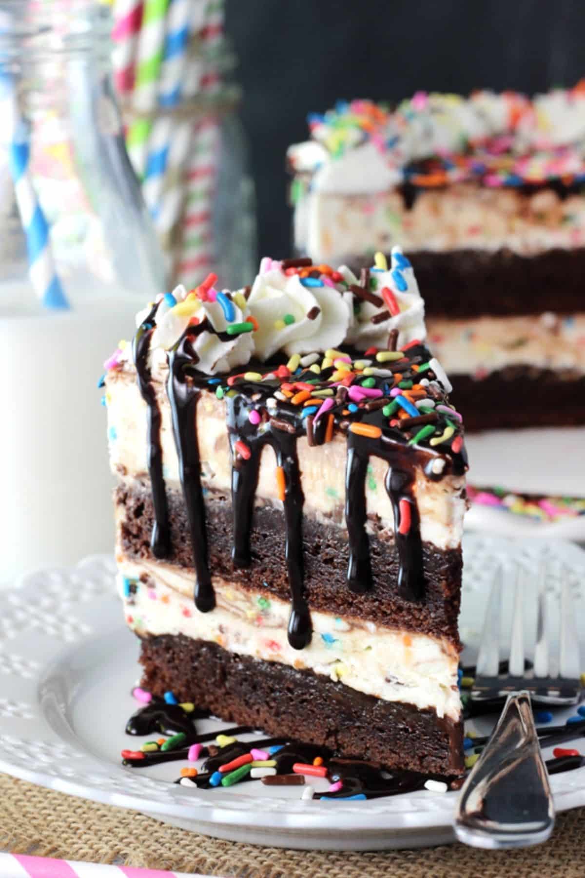 This Cake Batter Fudge Brownie Ice Cream Cake combines funfetti cake batter, brownies, fudge sauce and ice cream into one dessert! A slice of cake batter fudge brownie ice cream cake on a white plate with the rest of the cake in the background.