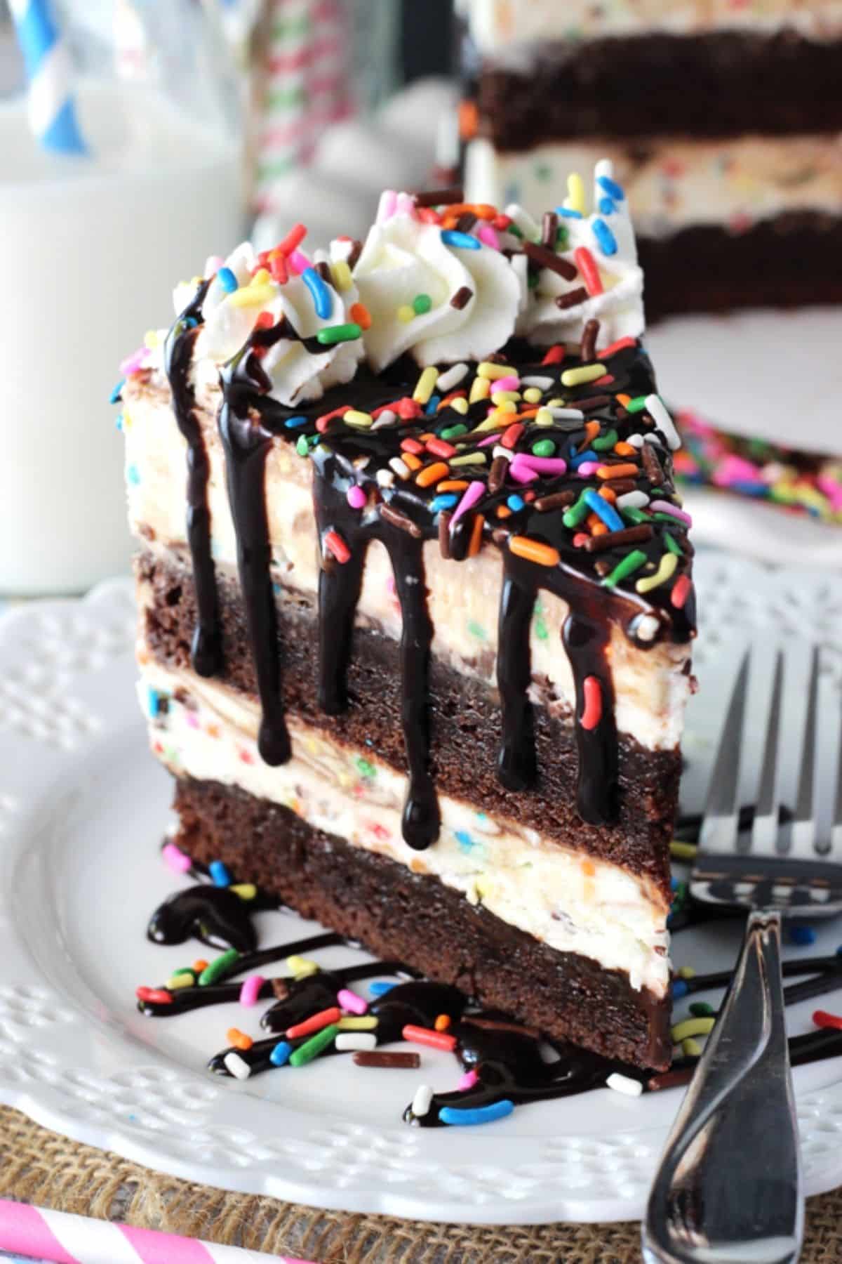 This Cake Batter Fudge Brownie Ice Cream Cake combines funfetti cake batter, brownies, fudge sauce and ice cream into one dessert! Side view of a slice of cake batter fudge brownie ice cream cake on a white plate next to a fork.