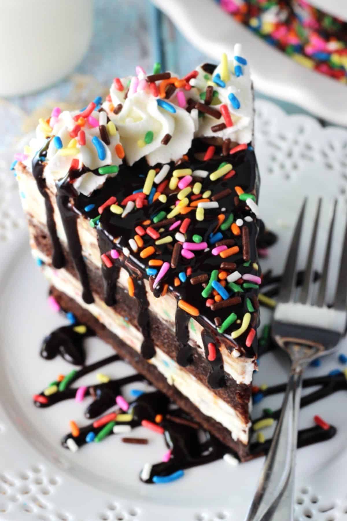 This Cake Batter Fudge Brownie Ice Cream Cake combines funfetti cake batter, brownies, fudge sauce and ice cream into one dessert! Top view of a slice of cake batter fudge brownie ice cream cake on a small white plate next to a fork.