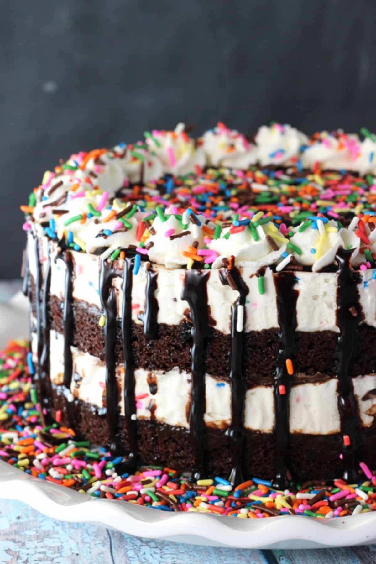 This Cake Batter Fudge Brownie Ice Cream Cake combines funfetti cake batter, brownies, fudge sauce and ice cream into one dessert! Close up of a cake batter fudge brownie ice cream cake on a white platter with lots of sprinkles.