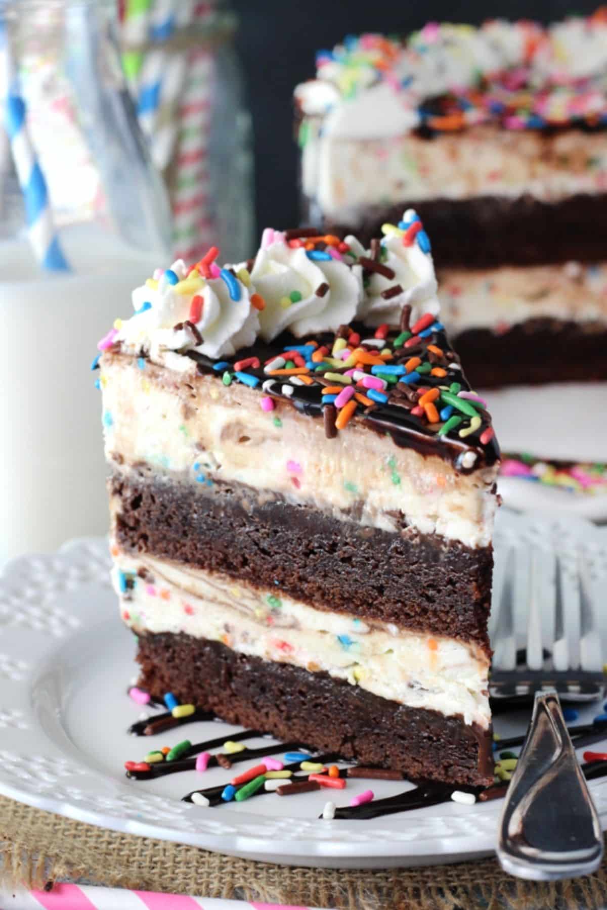 This Cake Batter Fudge Brownie Ice Cream Cake combines funfetti cake batter, brownies, fudge sauce and ice cream into one dessert! A slice of cake batter fudge brownie ice cream cake on a small white plate next to a fork.