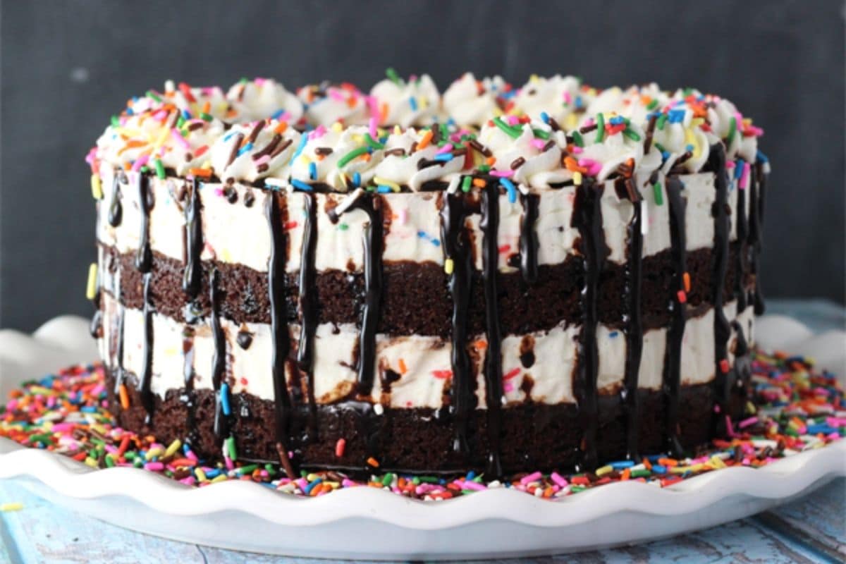 This Cake Batter Fudge Brownie Ice Cream Cake combines funfetti cake batter, brownies, fudge sauce and ice cream into one dessert! Close up of a cake batter fudge brownie ice cream cake on a white platter with lots of sprinkles.
