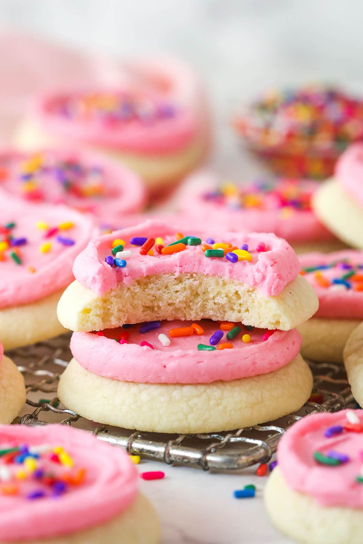 These copycat Lofthouse Cookies are soft and cakey with a melt-in-your-mouth texture and light flavor, just like the ones at the supermarket. Two copycat Lofthouse cookies stacked on a wire rack, one with a bite missing