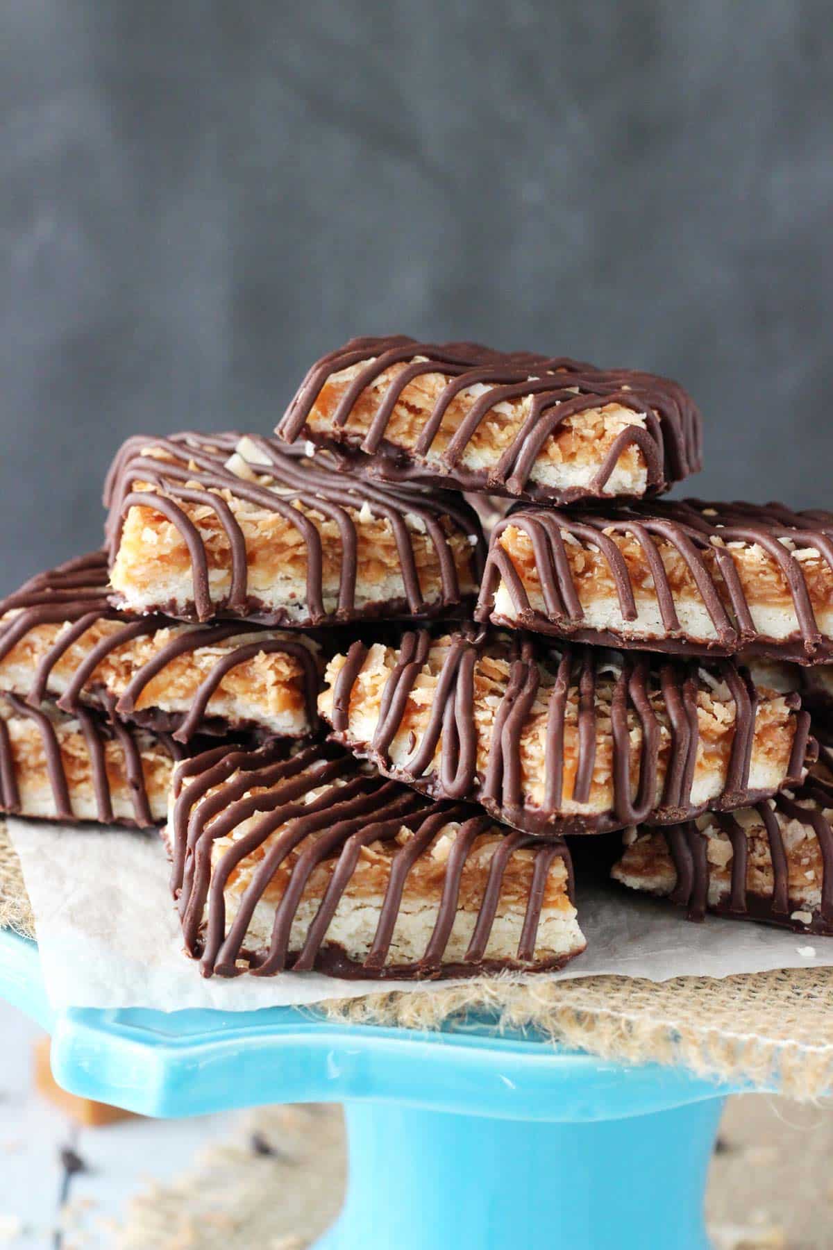 Buttery shortbread cookies are topped with gooey caramel, nutty toasted coconut and rich chocolate in these heavenly Samoa Cookie Bars! Close up of cut Samoa Cookie Bars on a blue stand.